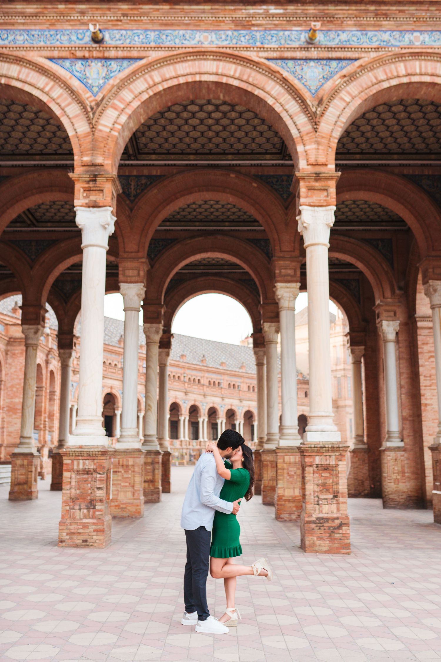 Couple embracing romantically under ornate brick arches at Plaza de España, Seville, Spain.