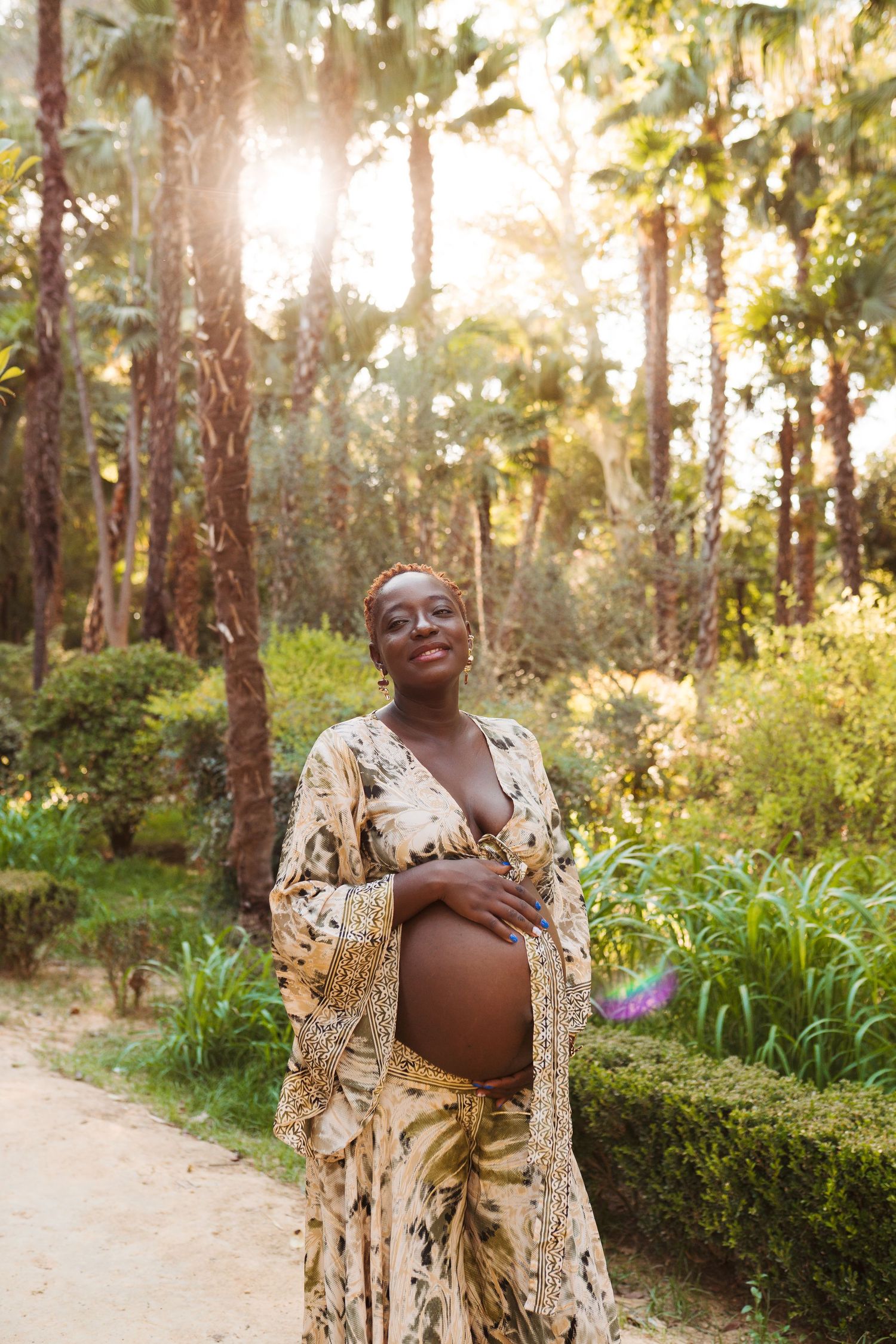 Pregnant woman in floral outfit cradling baby bump outdoors in sunlit tropical garden with palm trees.