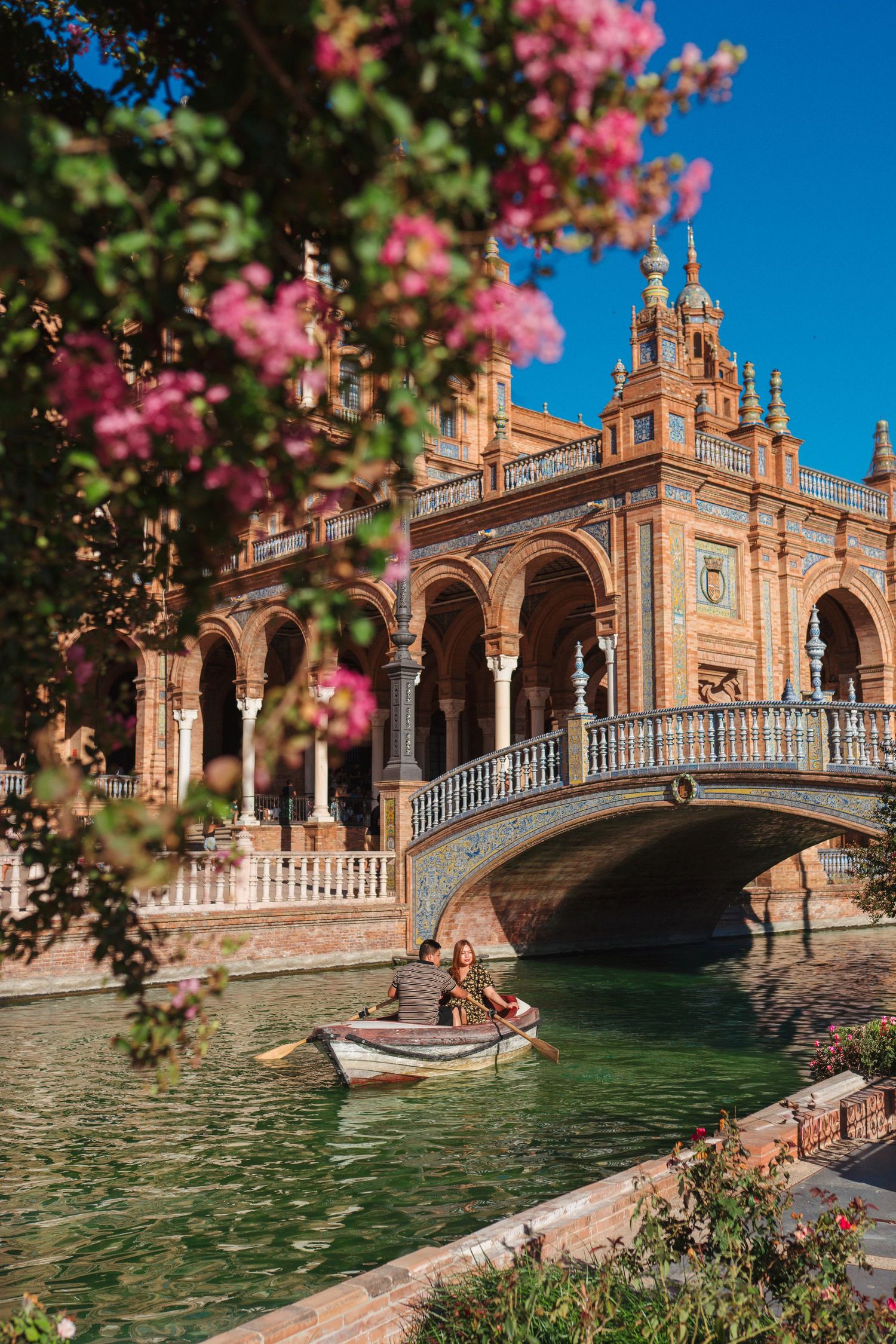 A scenic boat ride along the canal at Plaza de España in Seville, Spain, framed by pink flowers and ornate architecture.
