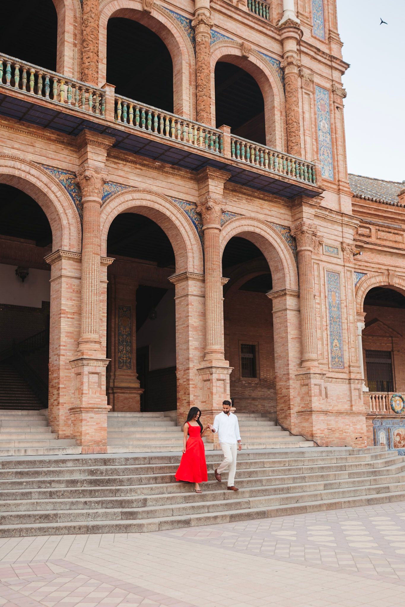 Couple in red dress and white shirt walking down grand stone steps beneath arched arcade at Plaza de España, Seville.
