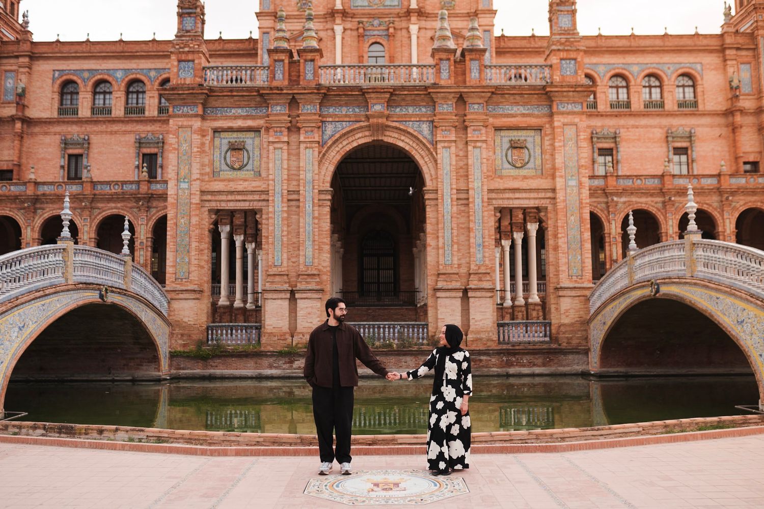 Couple holding hands at Plaza de España Seville with iconic bridge and canal in background.