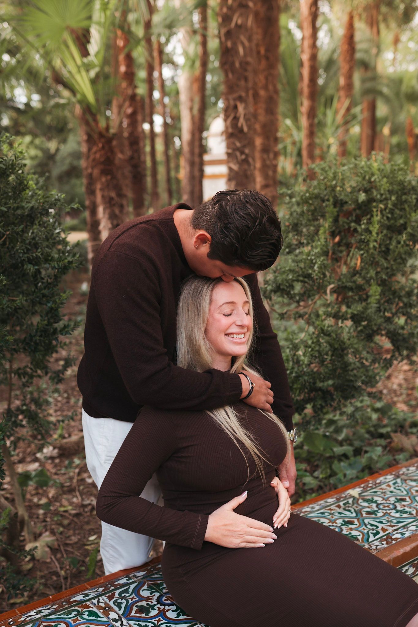 Smiling pregnant woman with partner embracing her among palm trees in park.