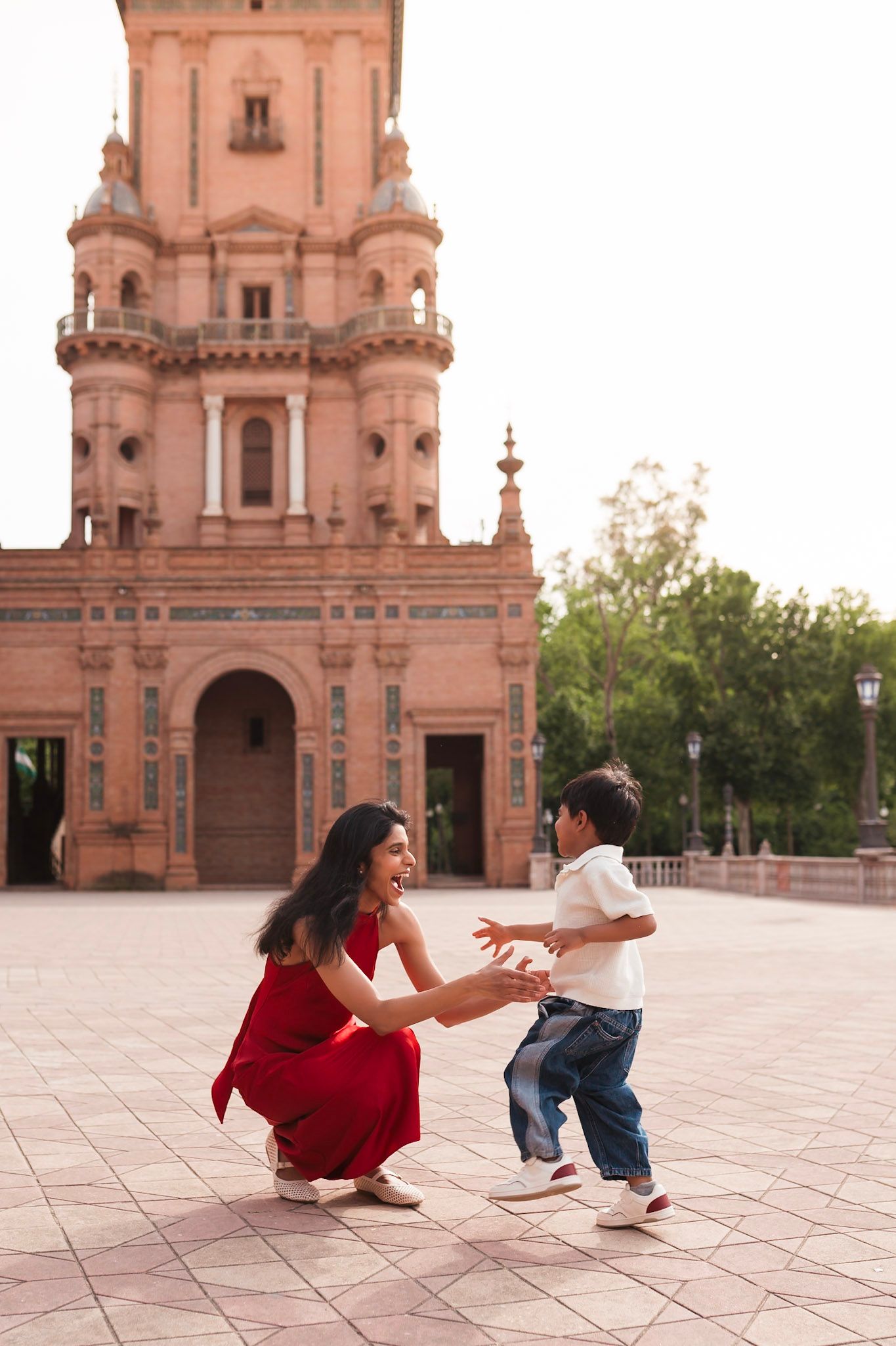 A mother in a red dress crouches to embrace her young son at Plaza de Espana in Seville Spain.