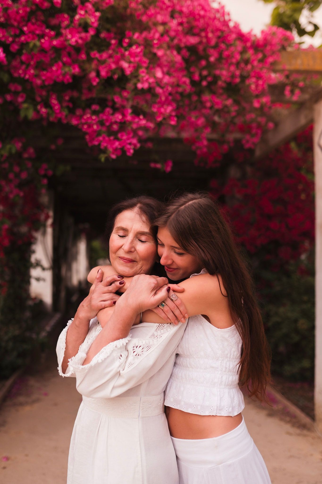 Two women in white dresses embracing warmly under vibrant pink bougainvillea flowers.