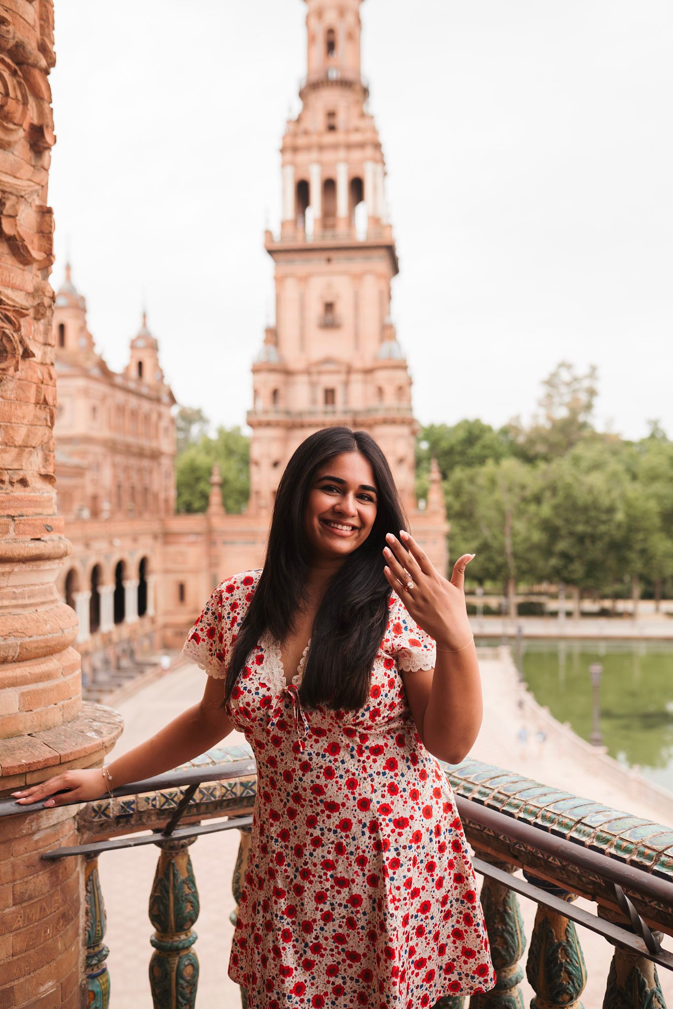 Smiling woman in red floral dress waving at Plaza de España, Seville, Spain.