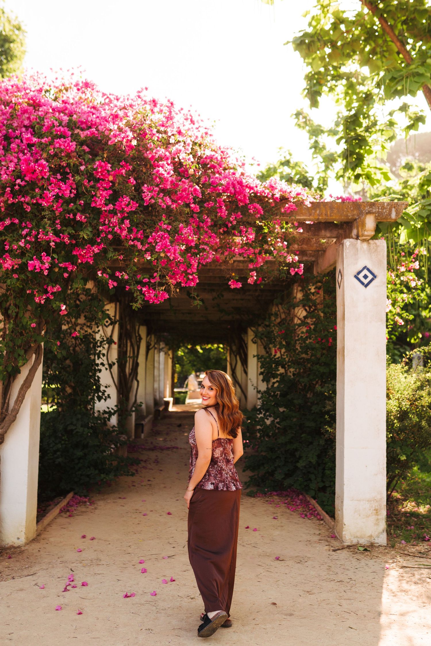 Woman in burgundy maxi dress poses under a pergola covered in vibrant pink bougainvillea flowers.
