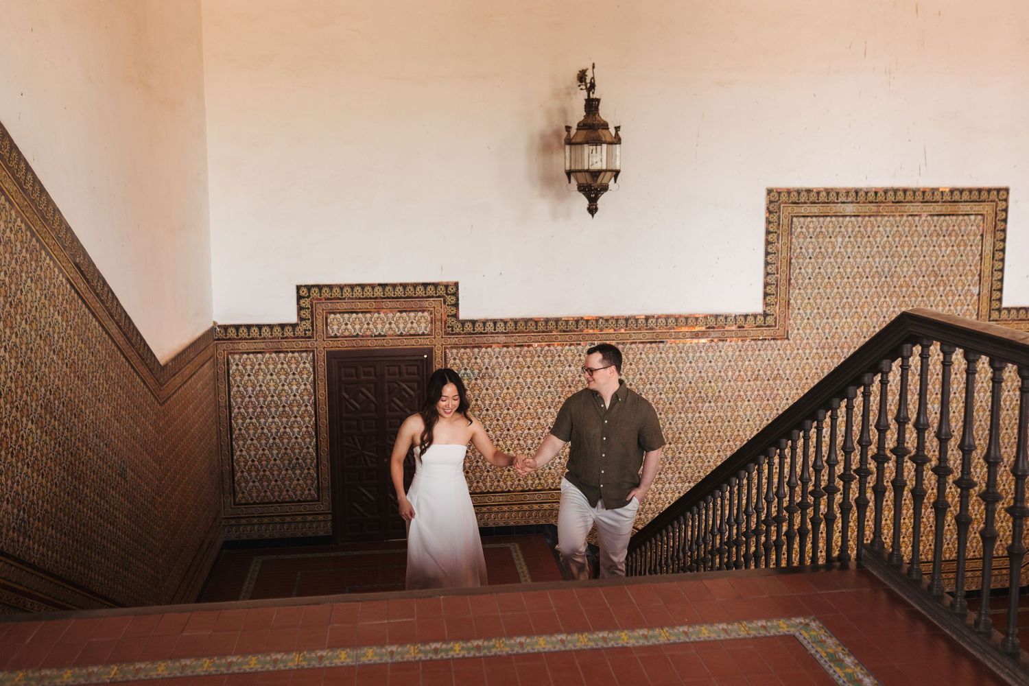 Couple holding hands on ornate tiled staircase in historic building, woman in white dress, man in green shirt.