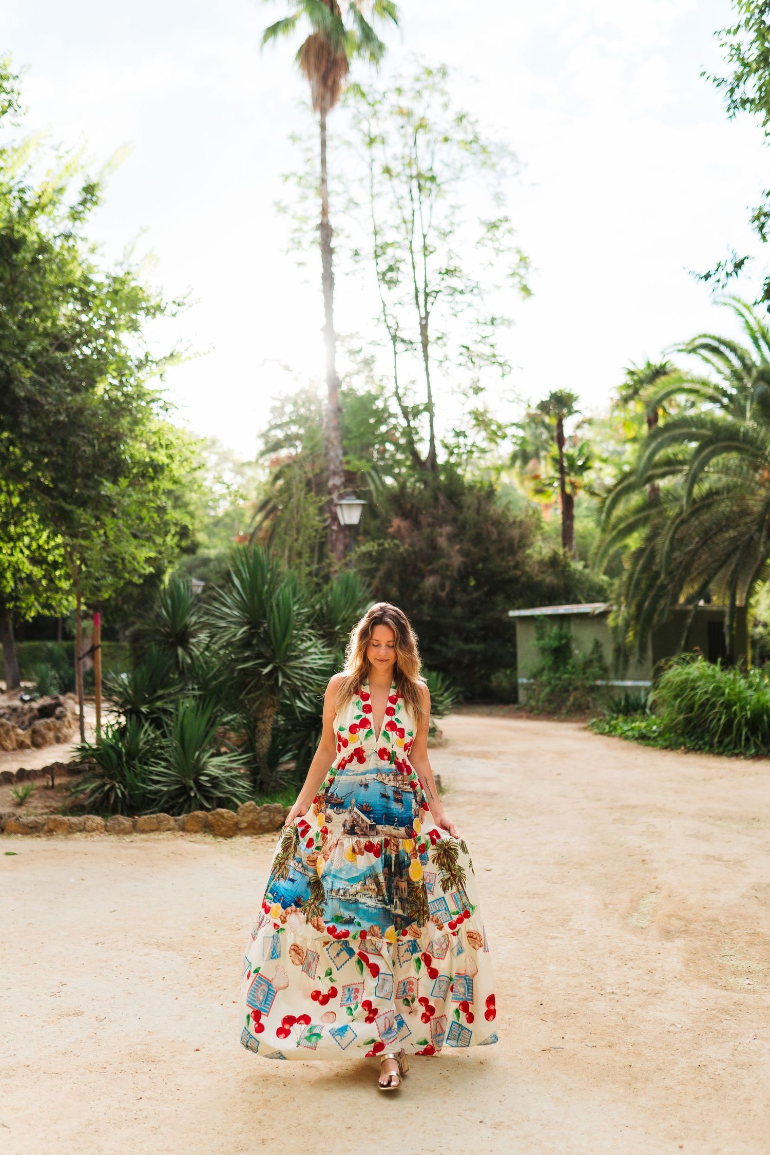 Woman in colorful floral maxi dress walking along a sunny park path surrounded by lush palm trees.