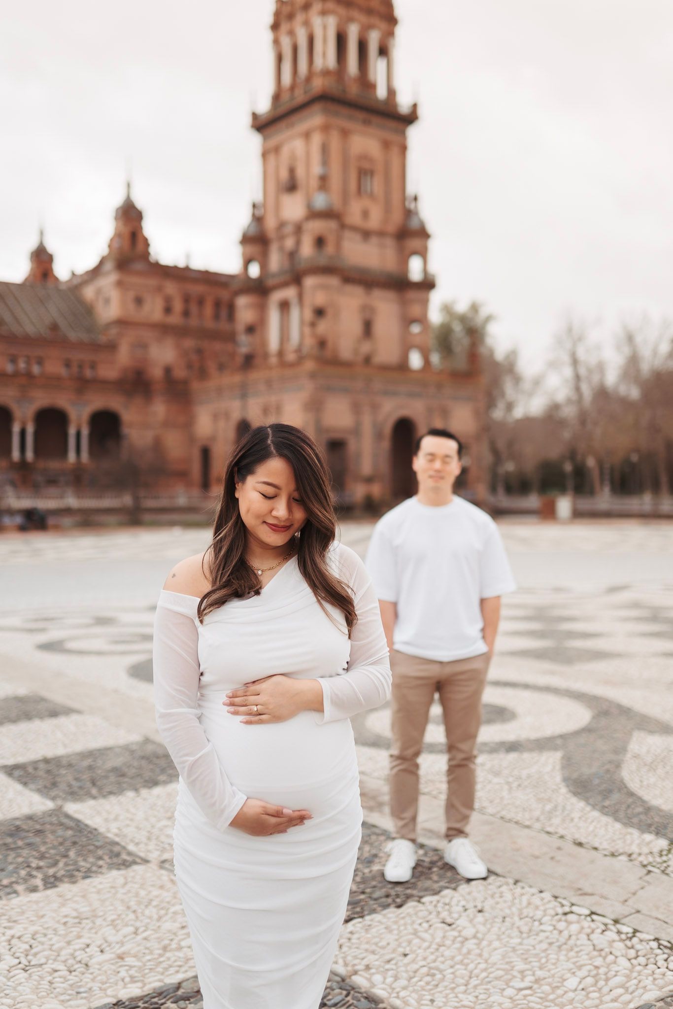 Pregnant woman in white dress holds baby bump as partner stands behind her at Plaza de Espana.