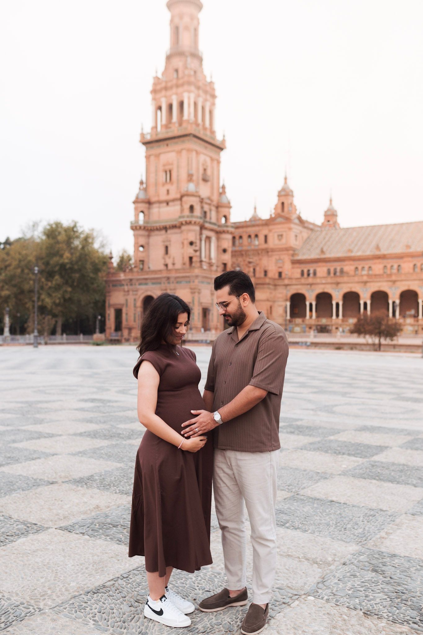 Expecting couple in brown outfits holding baby bump at Plaza de España, Seville.