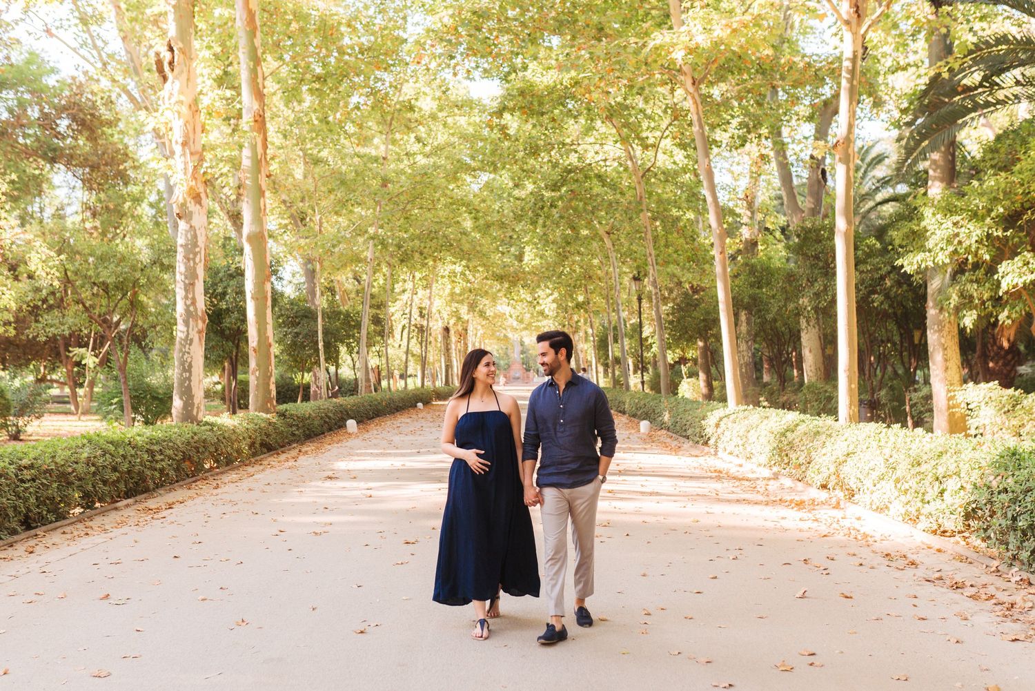 Pregnant couple walking hand in hand along a tree-lined park path, woman in navy dress, man in blue shirt.