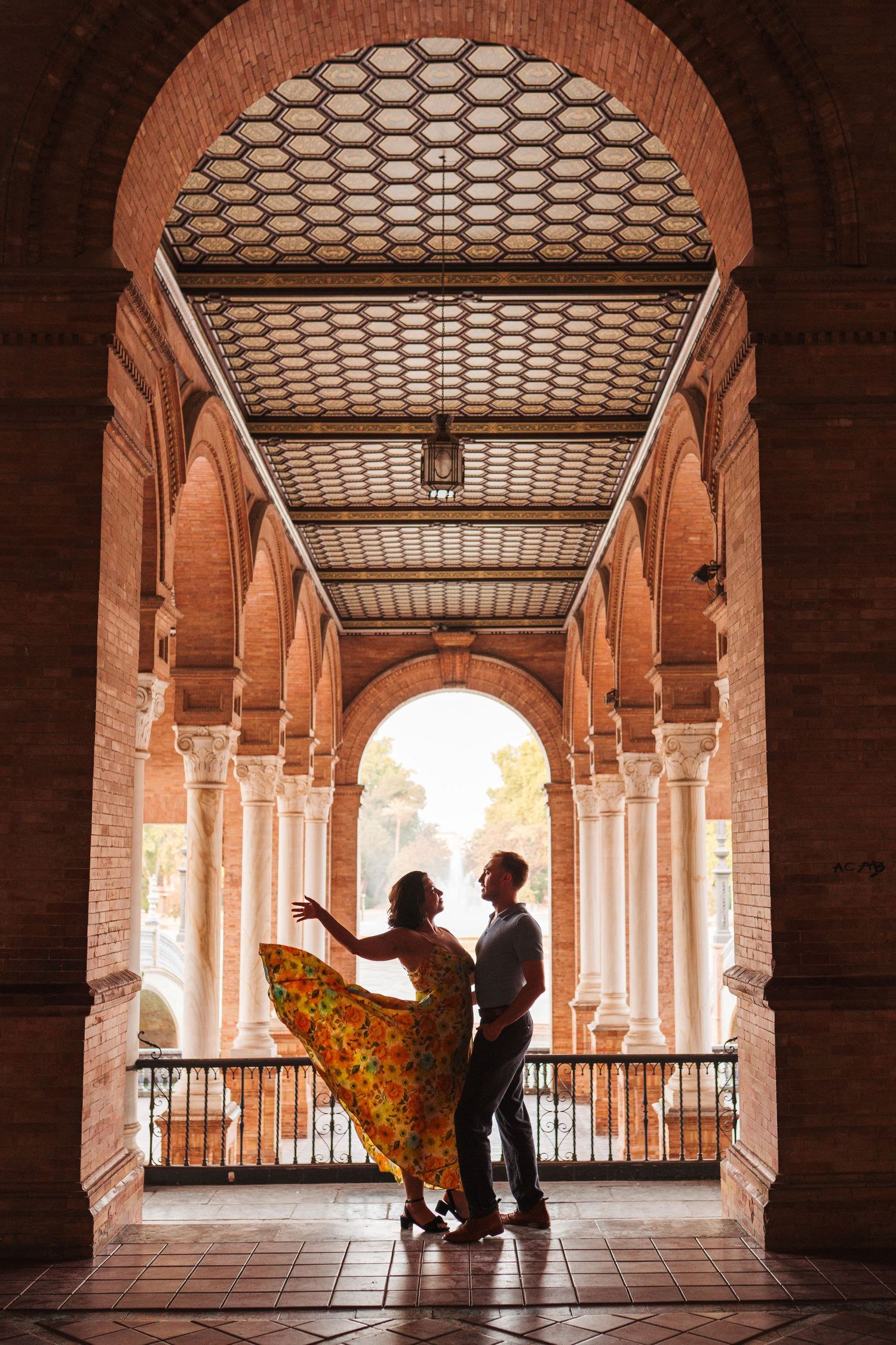 Couple dancing flamenco under ornate brick arches at Plaza de España, Seville, woman in flowing yellow dress.