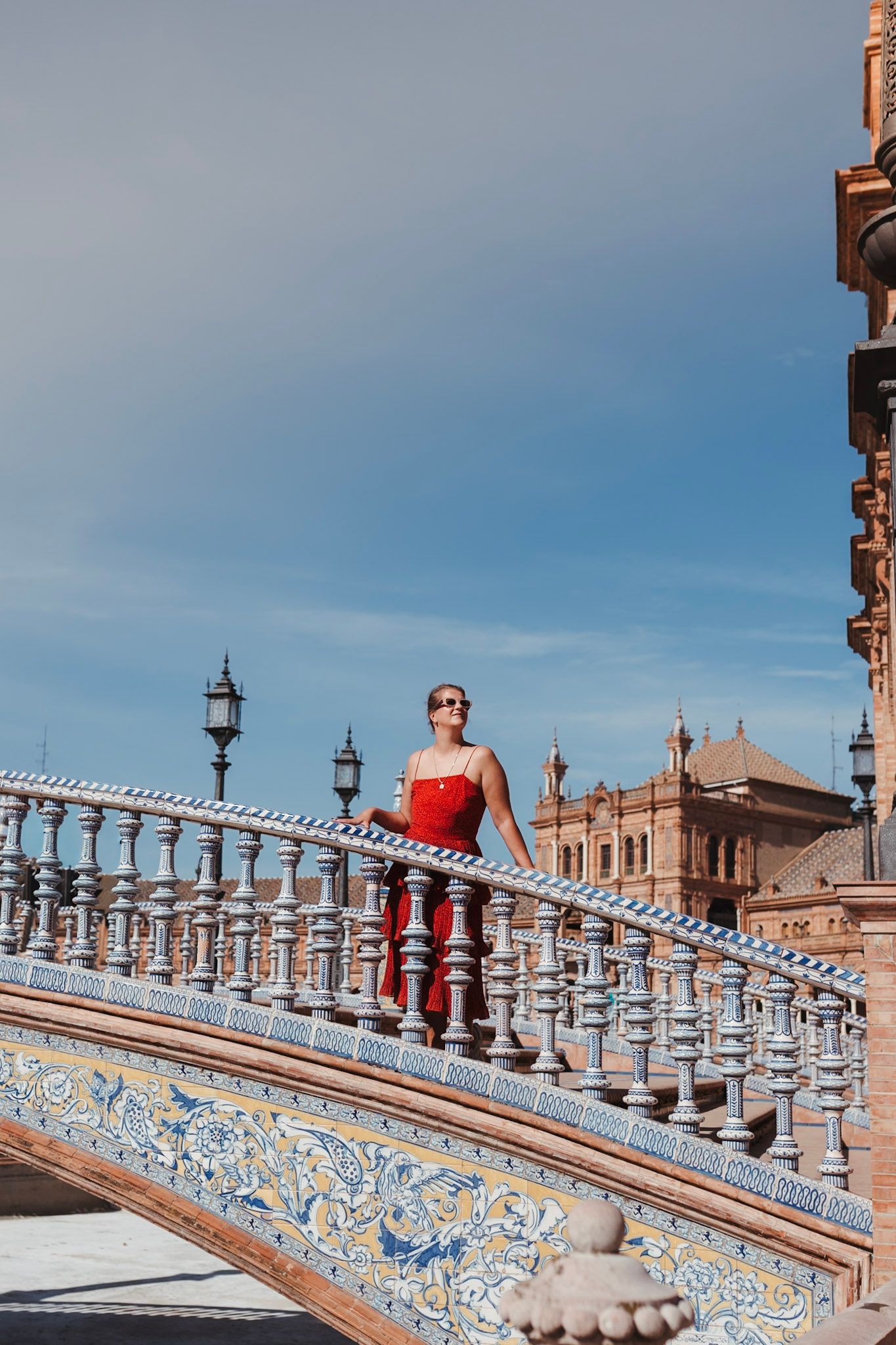 Woman in red dress standing on ornate tiled bridge at Plaza de España under blue sky in Seville.