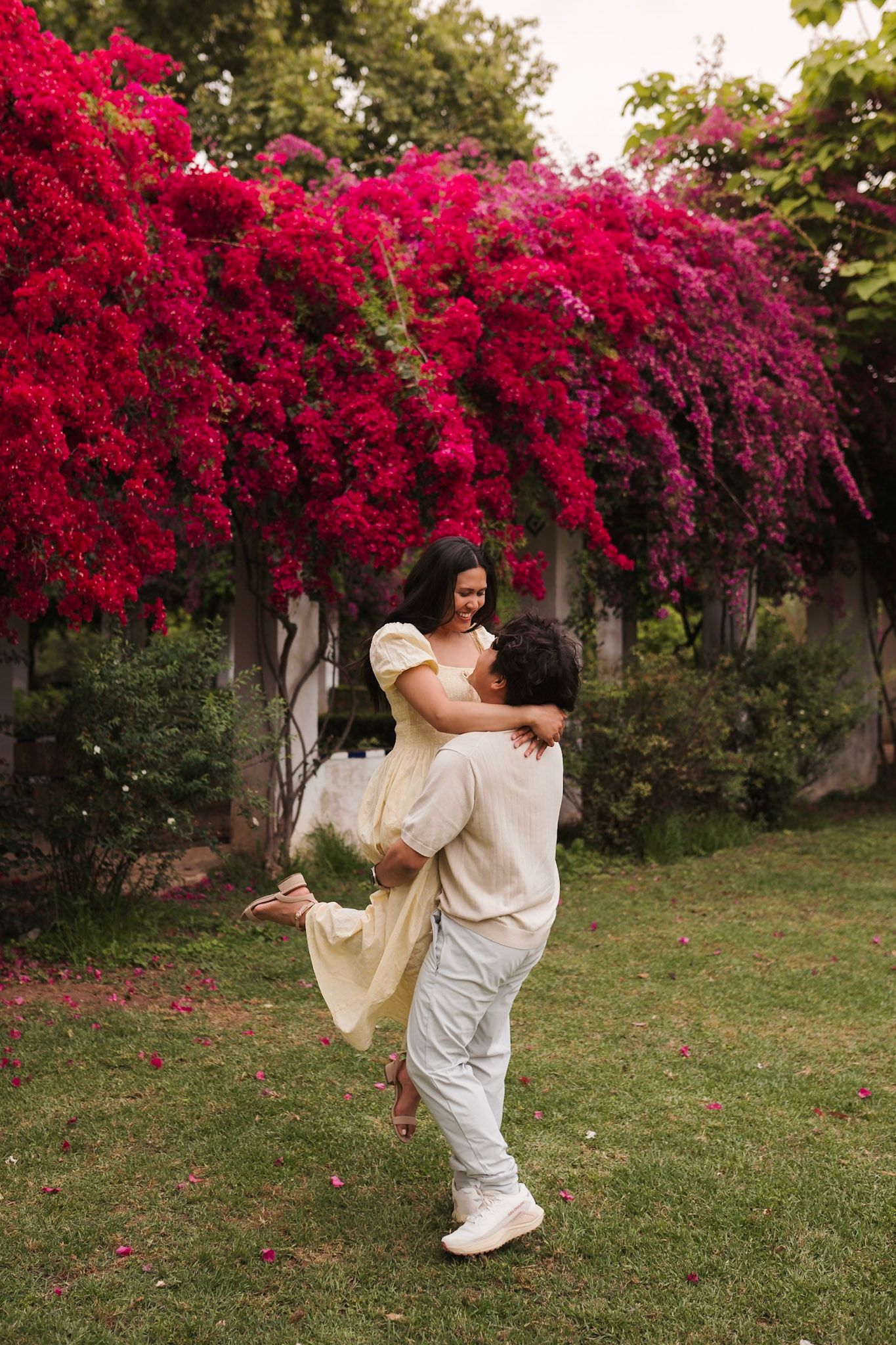 Couple embracing in front of vibrant red bougainvillea flowers in a lush garden.