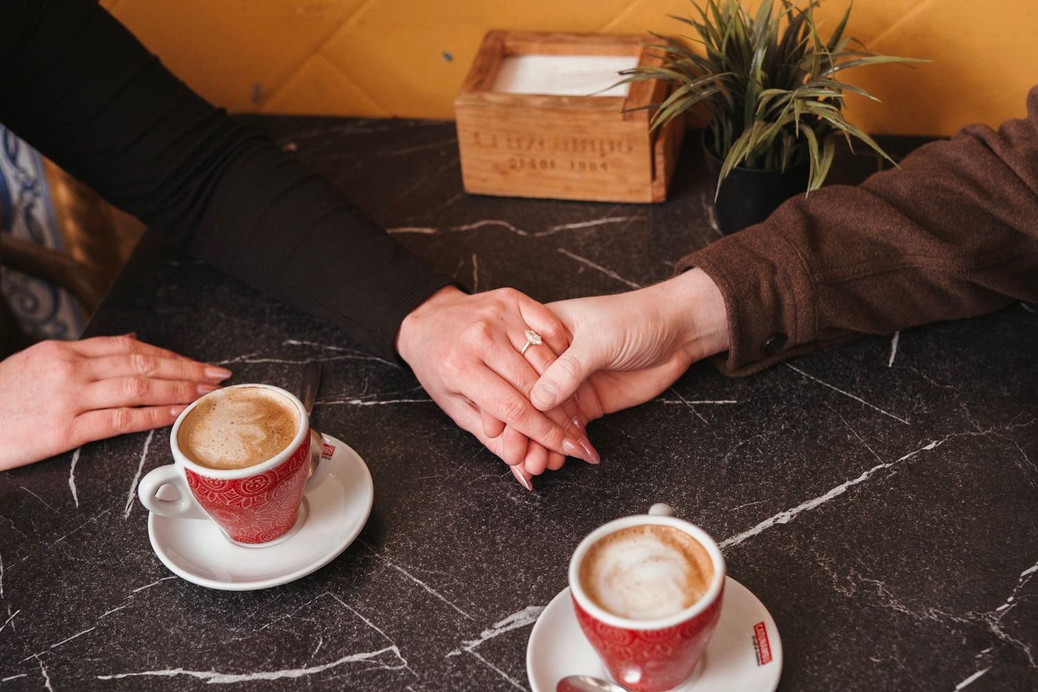 Engaged couple holding hands over marble table with two cappuccinos after proposal celebration.
