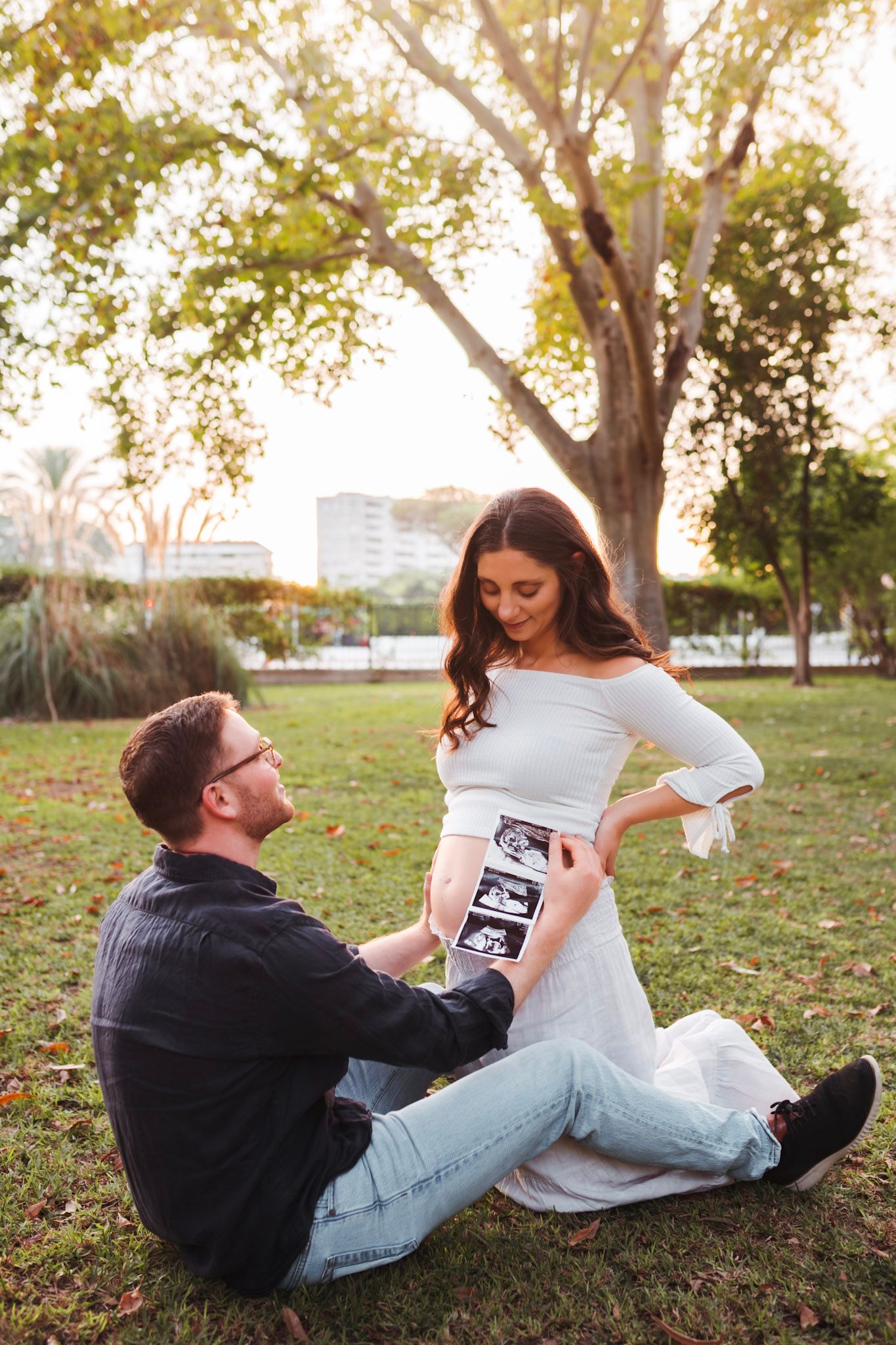 Man sitting on grass holds ultrasound photos near pregnant woman in white dress.