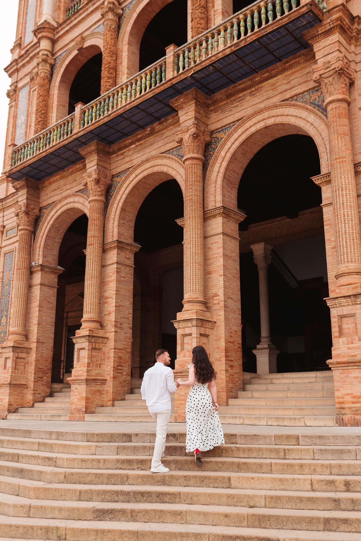 Couple holding hands walking up steps of ornate brick building with arched columns at Plaza de Espana, Seville.