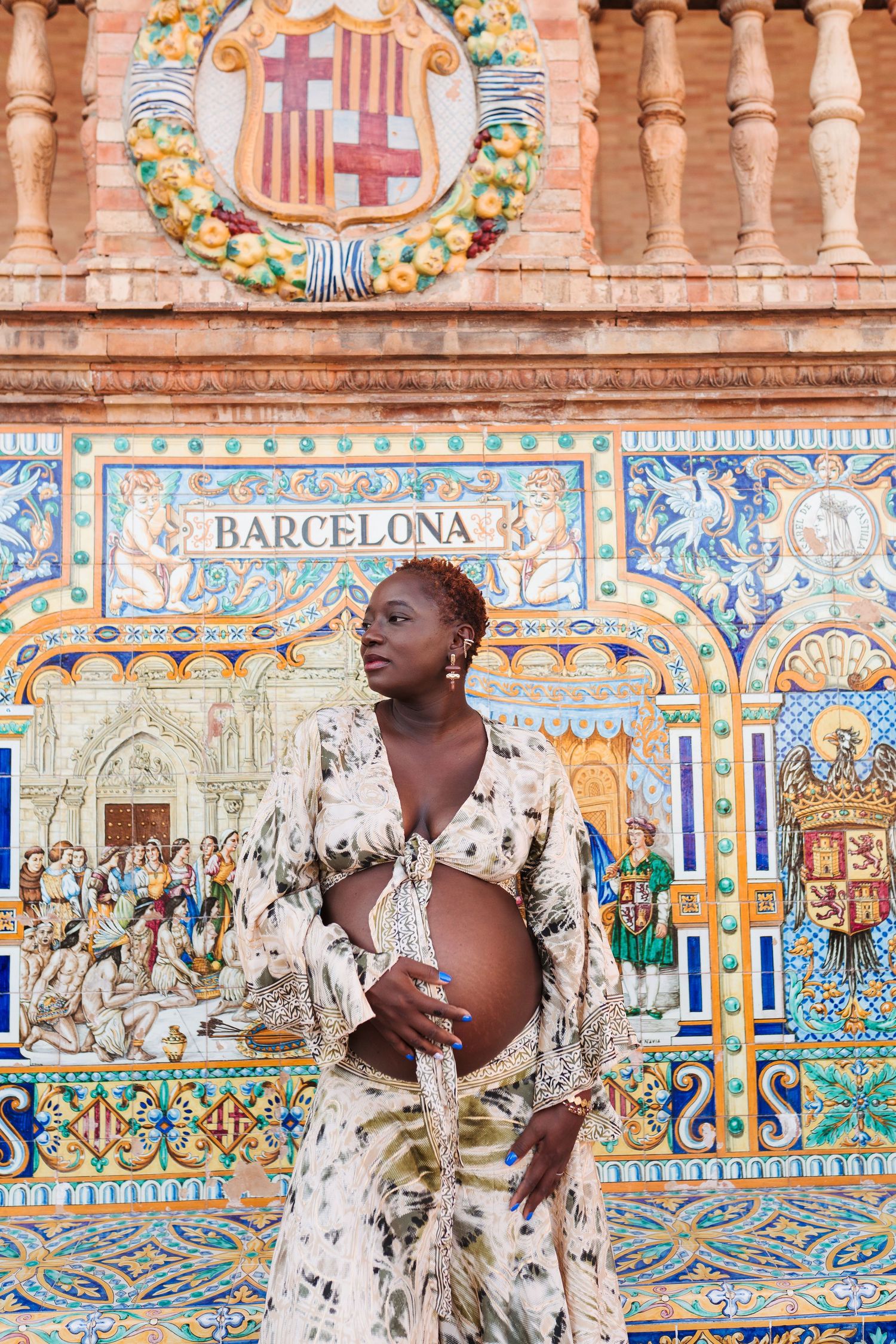 Pregnant woman in floral dress poses before Barcelona tile mosaic at Plaza de España, Seville.