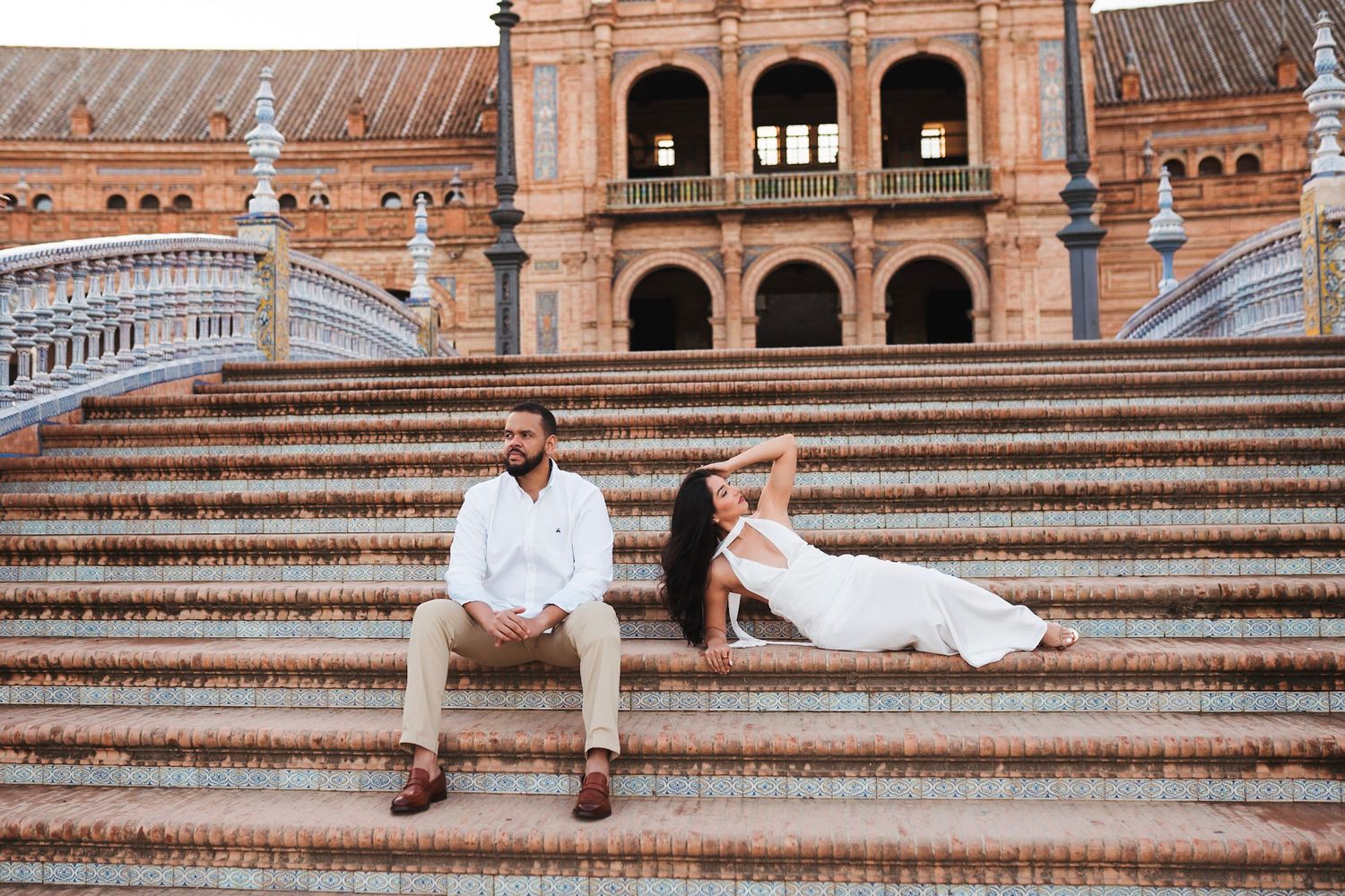 Couple sitting apart on grand steps of Plaza de Espana in Seville, woman in white dress reclining gracefully.