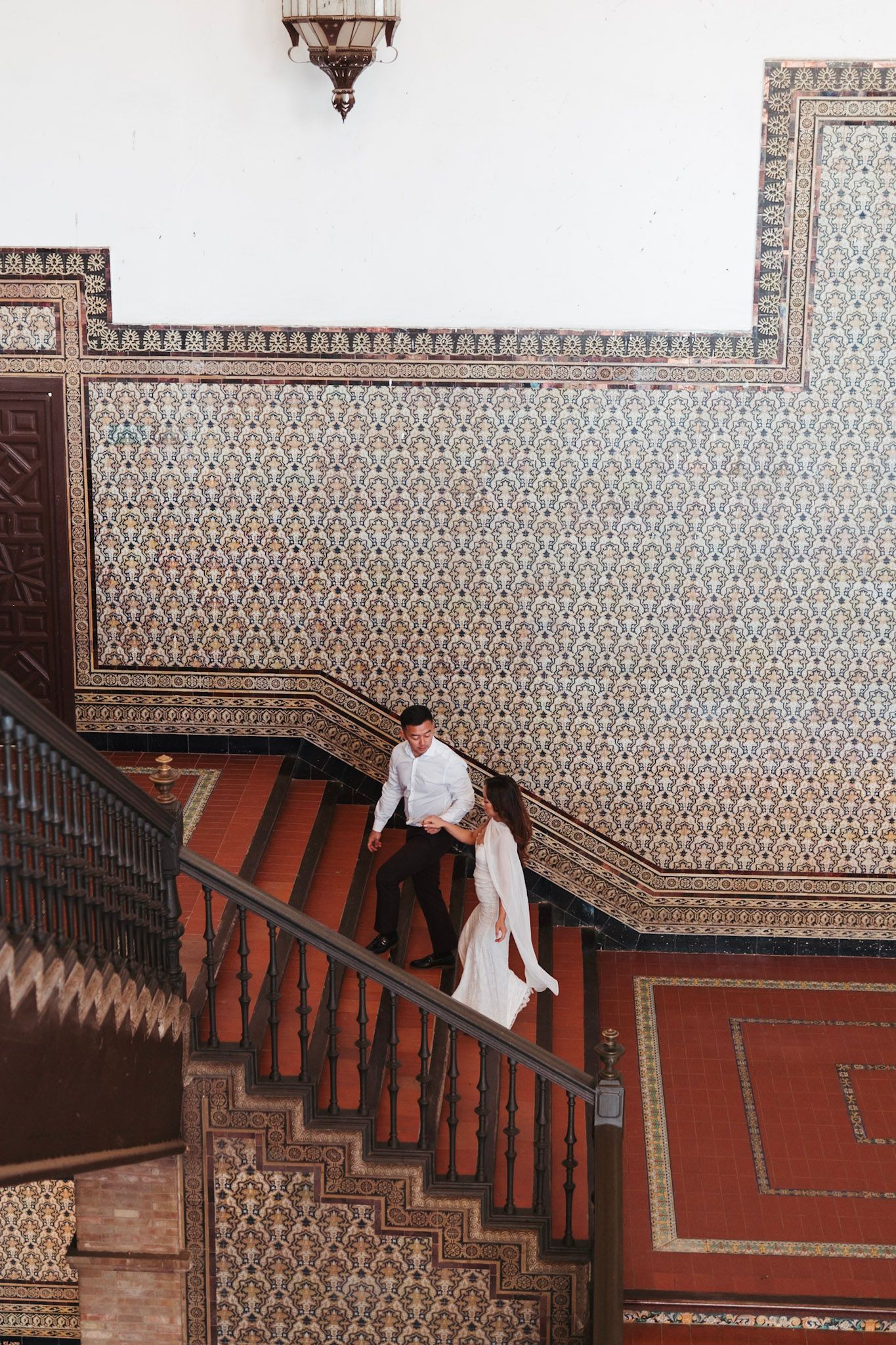 Couple descending ornate Moorish tiled staircase in a historic building in Seville, Spain.