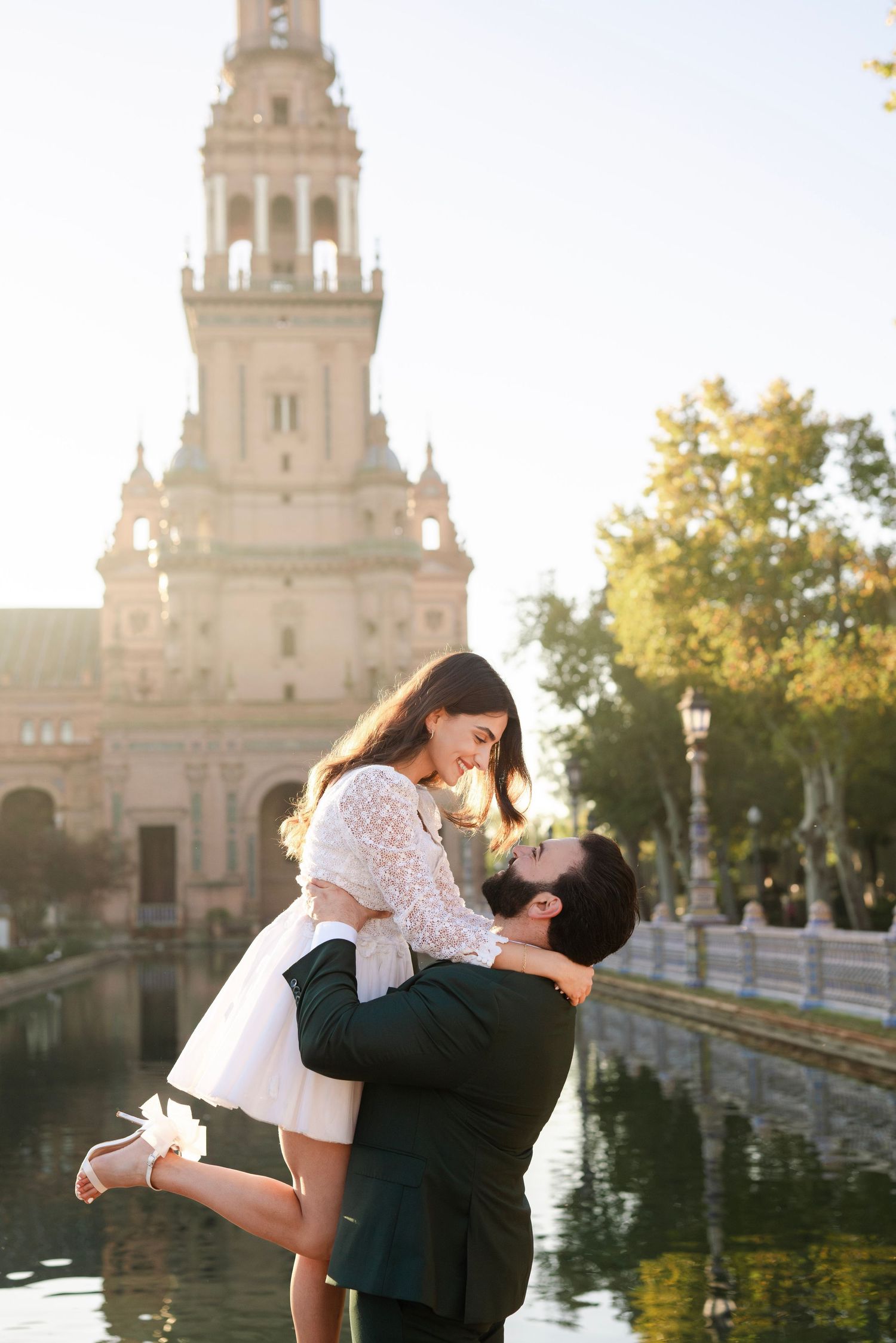 Man lifting woman near a canal at golden hour with Plaza de España tower glowing in the background.