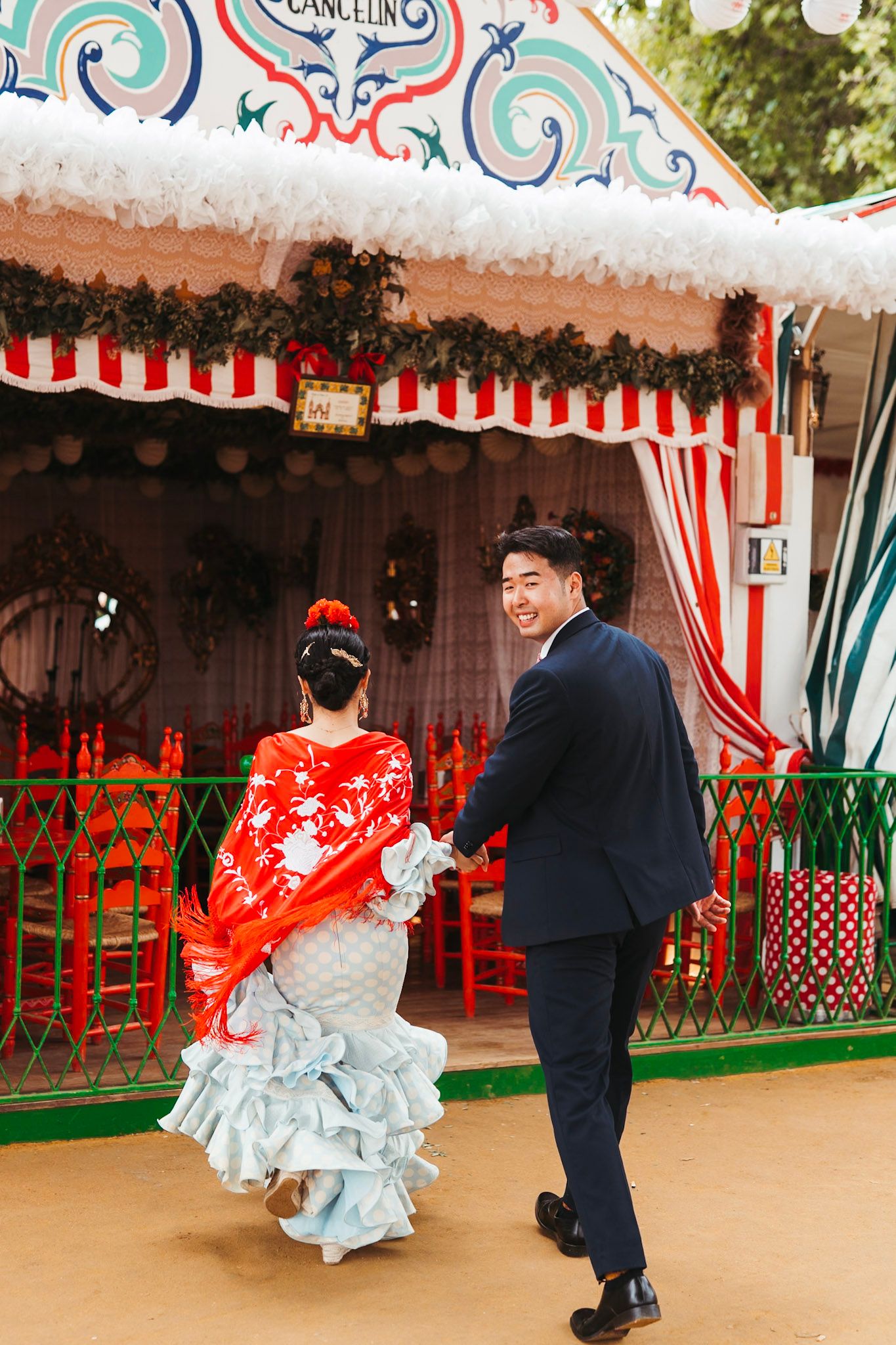 Smiling couple in flamenco dress and suit walk hand in hand at Seville Fair.