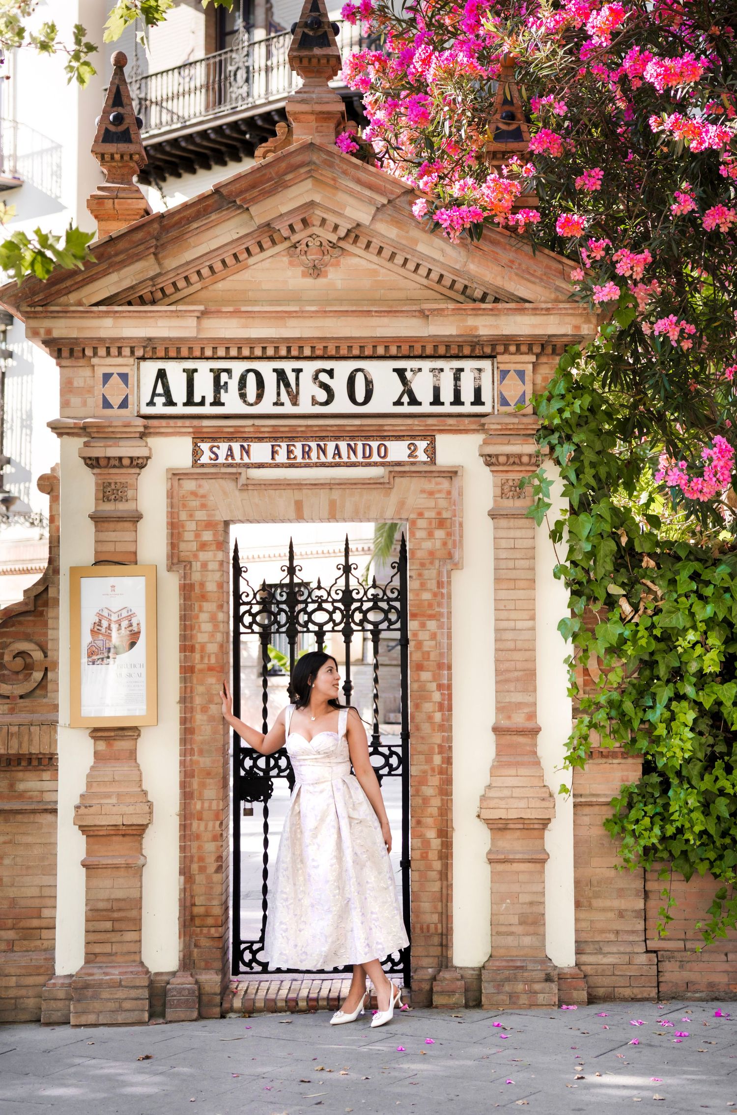 Woman in white dress stands at ornate Alfonso XIII gate on San Fernando street in Seville, Spain.
