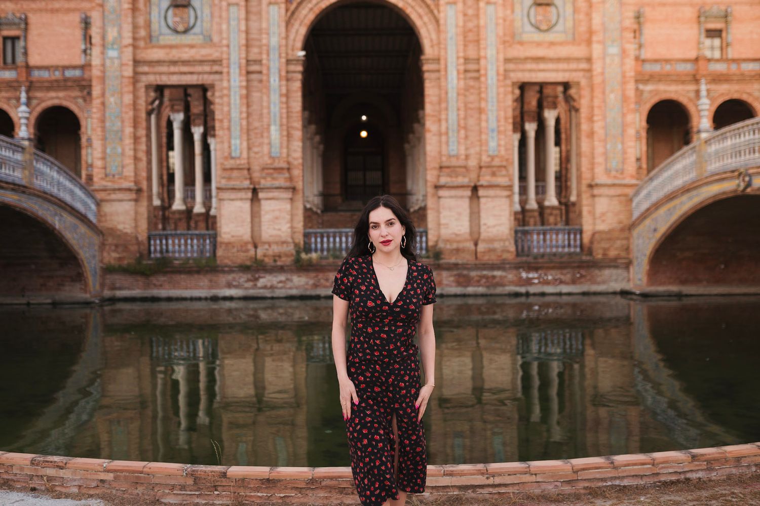 Woman in black floral dress stands before arched Plaza de España reflecting pool.