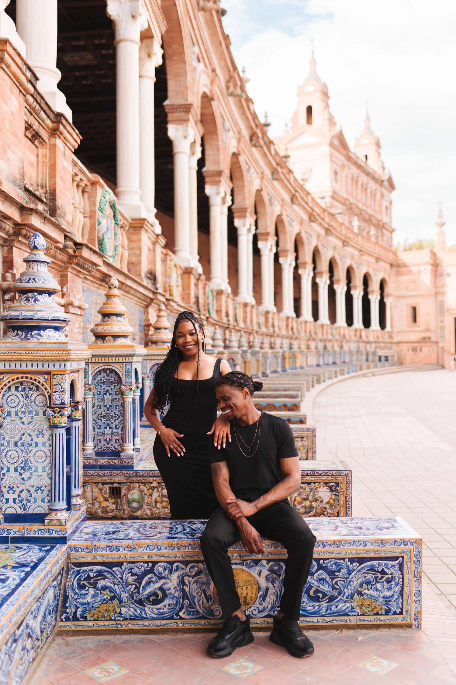 Couple in black outfits posing at Plaza de España, Seville, with colorful tiled benches and ornate arches.