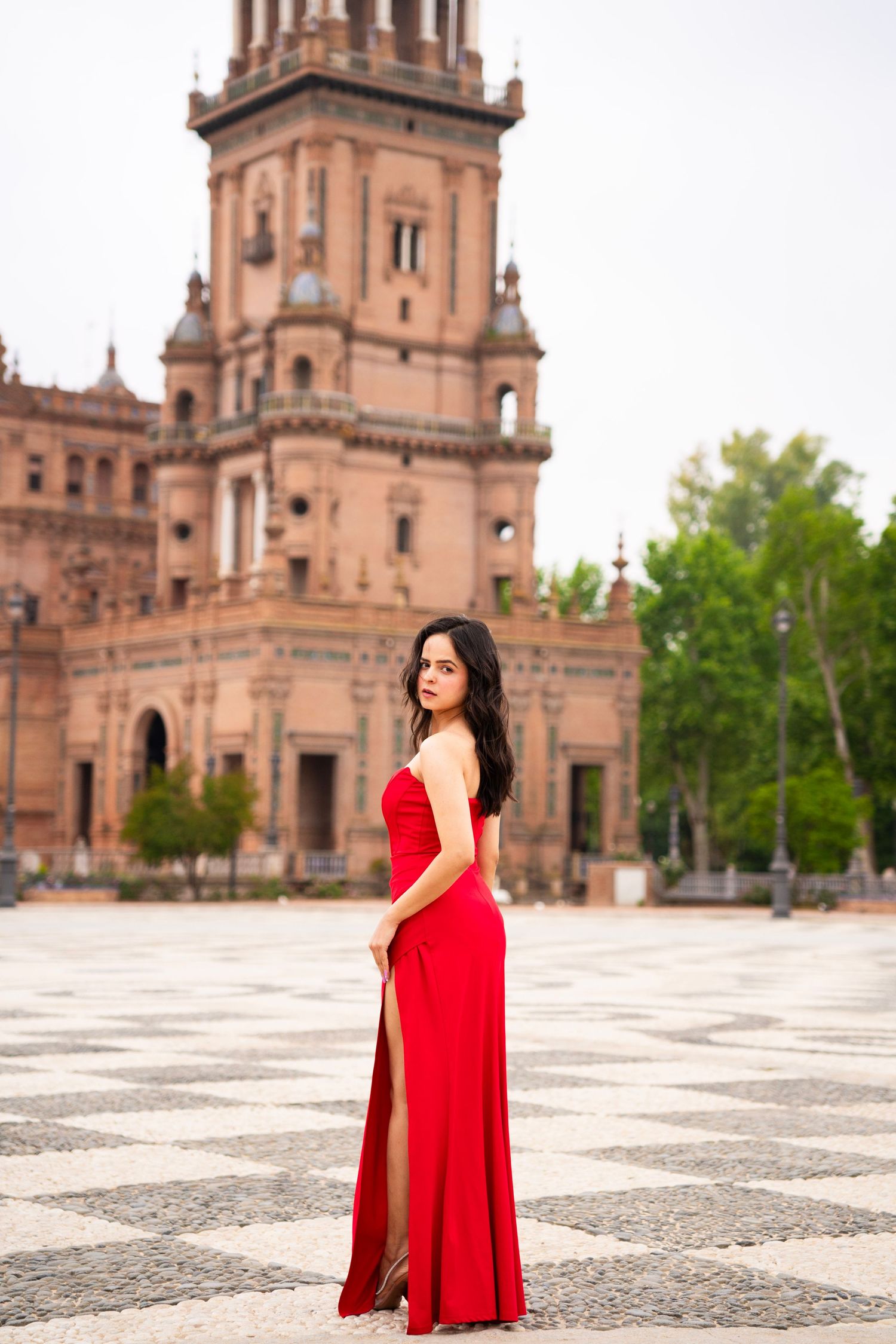 Woman in elegant red slit gown stands on checkered plaza floor before historic Seville tower landmark.