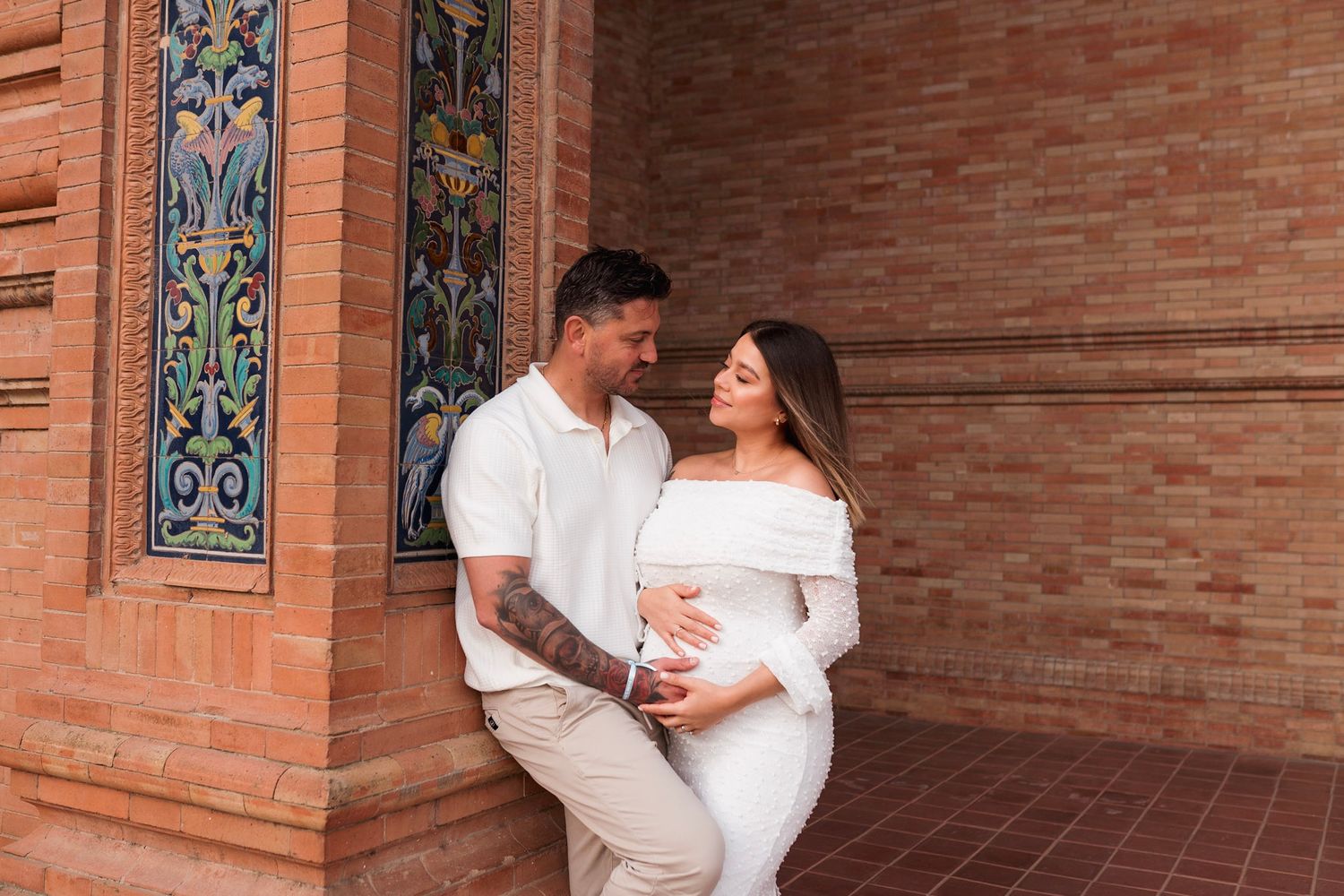 Expecting couple in white outfits embrace beside ornate tiled wall at Plaza de España Seville.