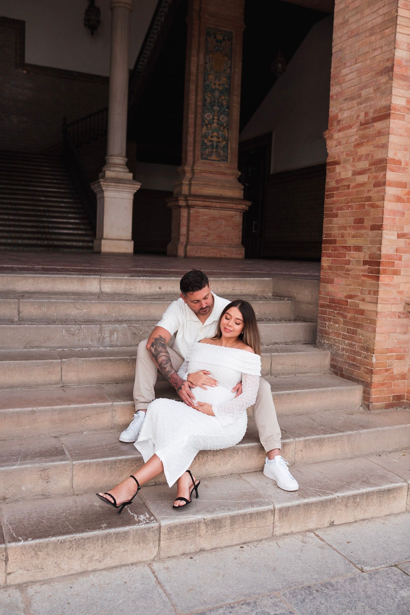 Couple sitting on steps at Plaza de España Seville, man tenderly holds pregnant womans belly.