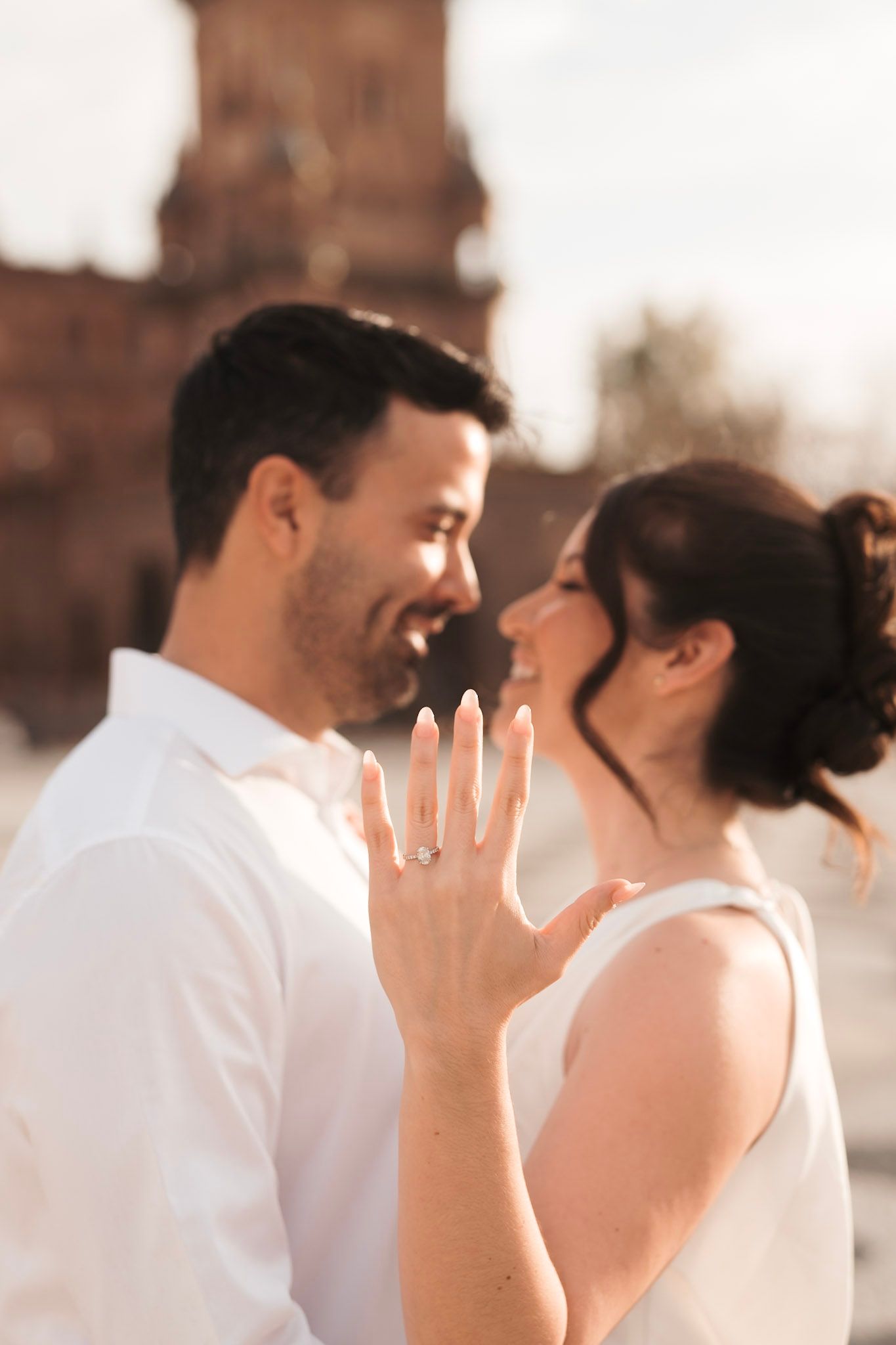 Happy couple nearly kissing at Plaza de España, woman displaying diamond engagement ring toward camera.