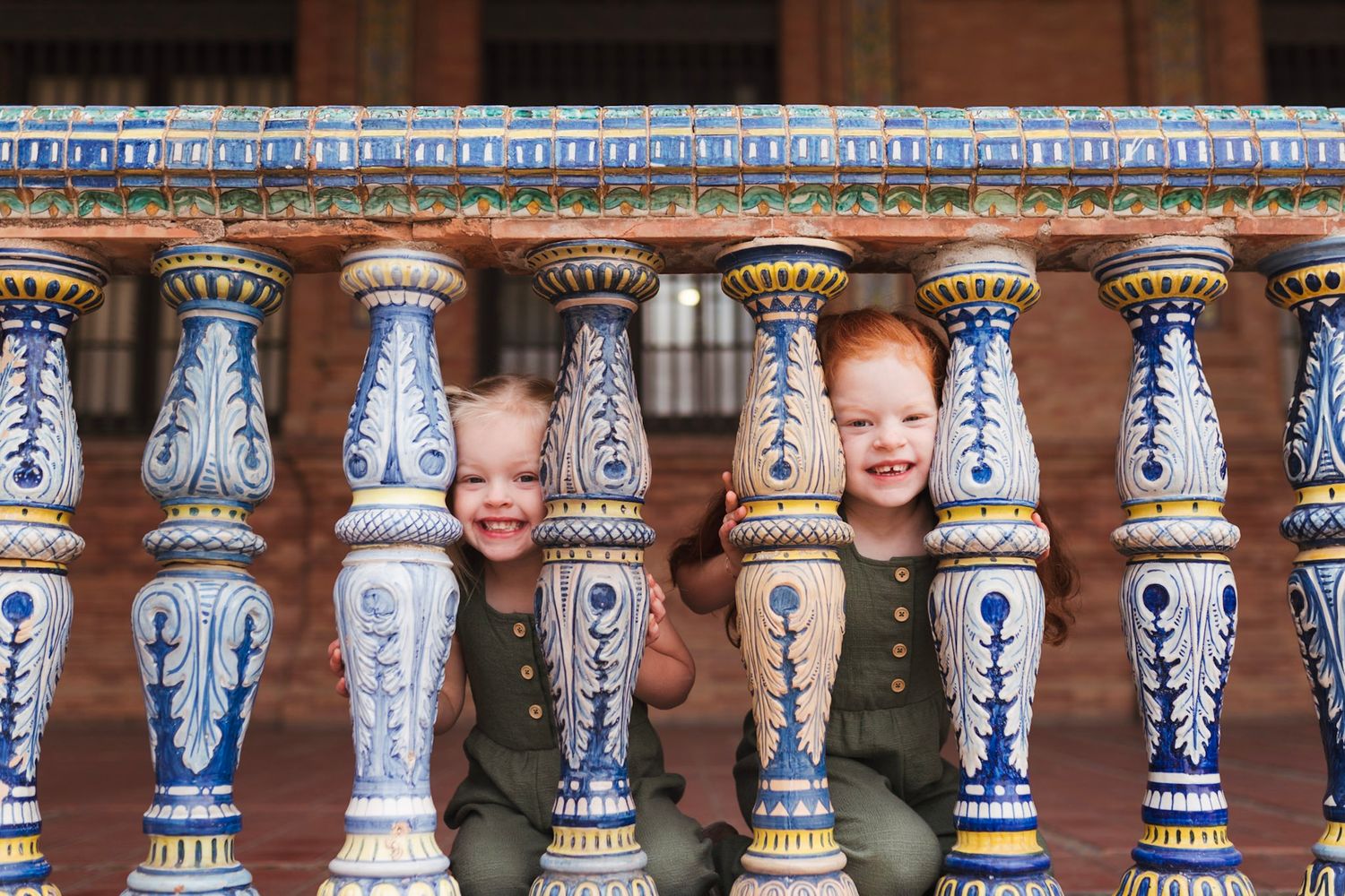 Two adorable red-haired toddlers smiling through ornate blue and white ceramic railings in Seville.
