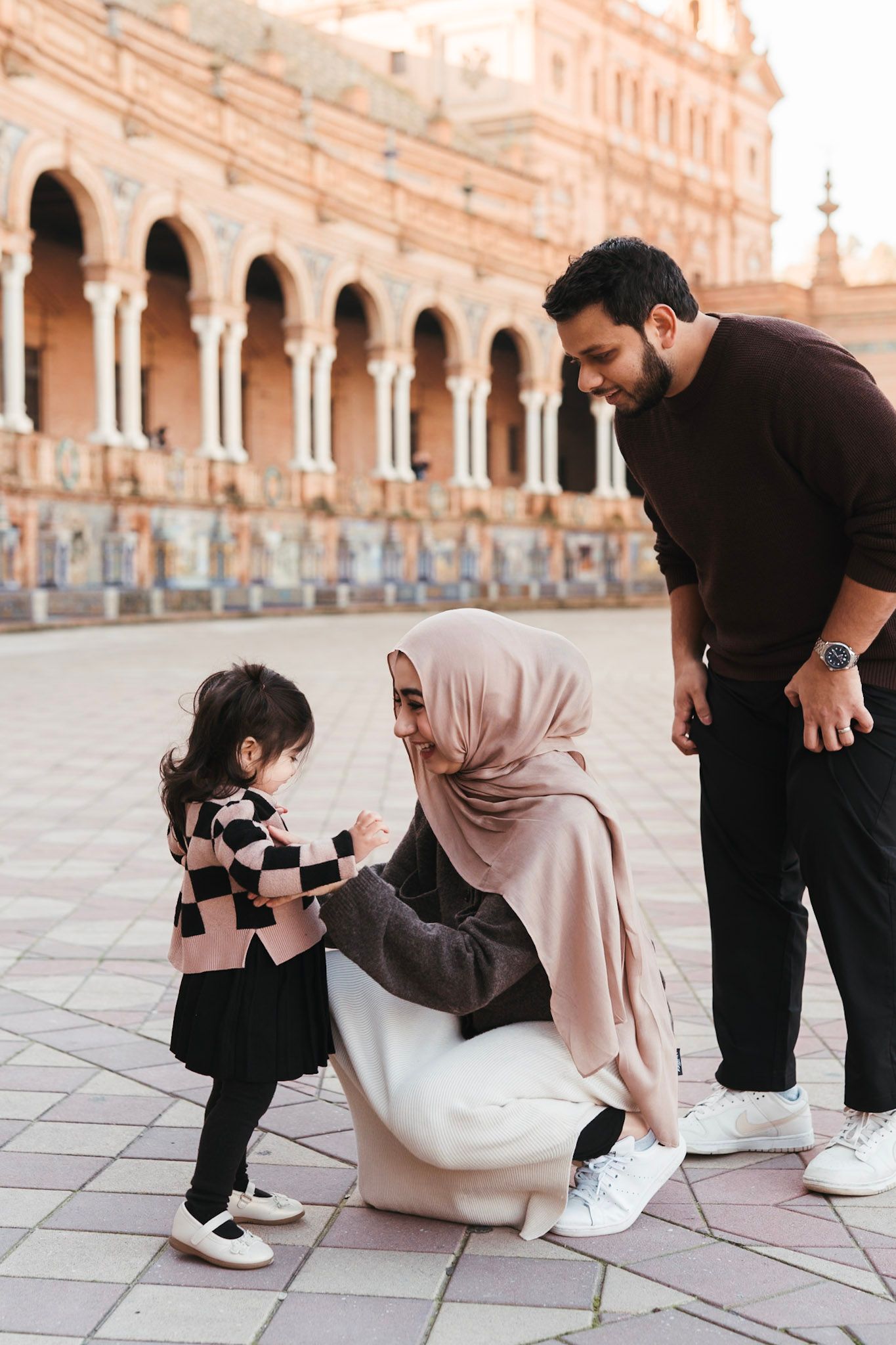 A Muslim family with a young daughter interacts warmly at Plaza de España, Seville, at golden hour.