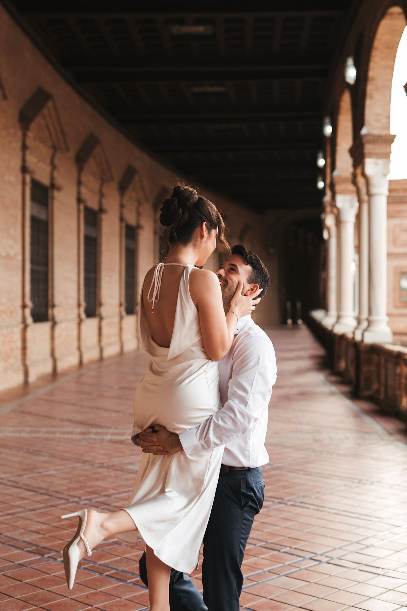 Man lifting woman in white dress for a kiss in a brick archway corridor.