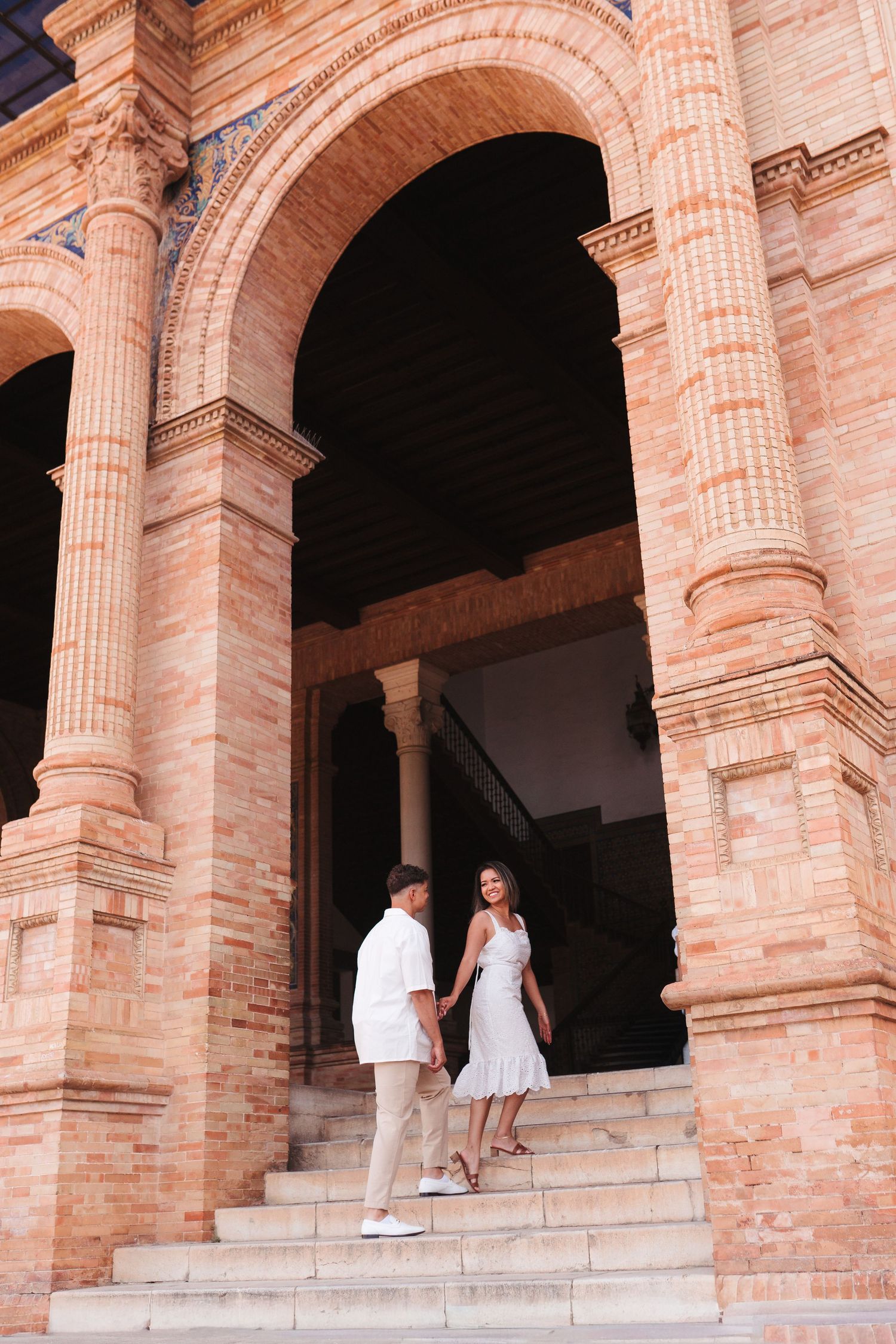 Couple holding hands on steps of ornate brick archway building in Seville, Spain, wearing white outfits.