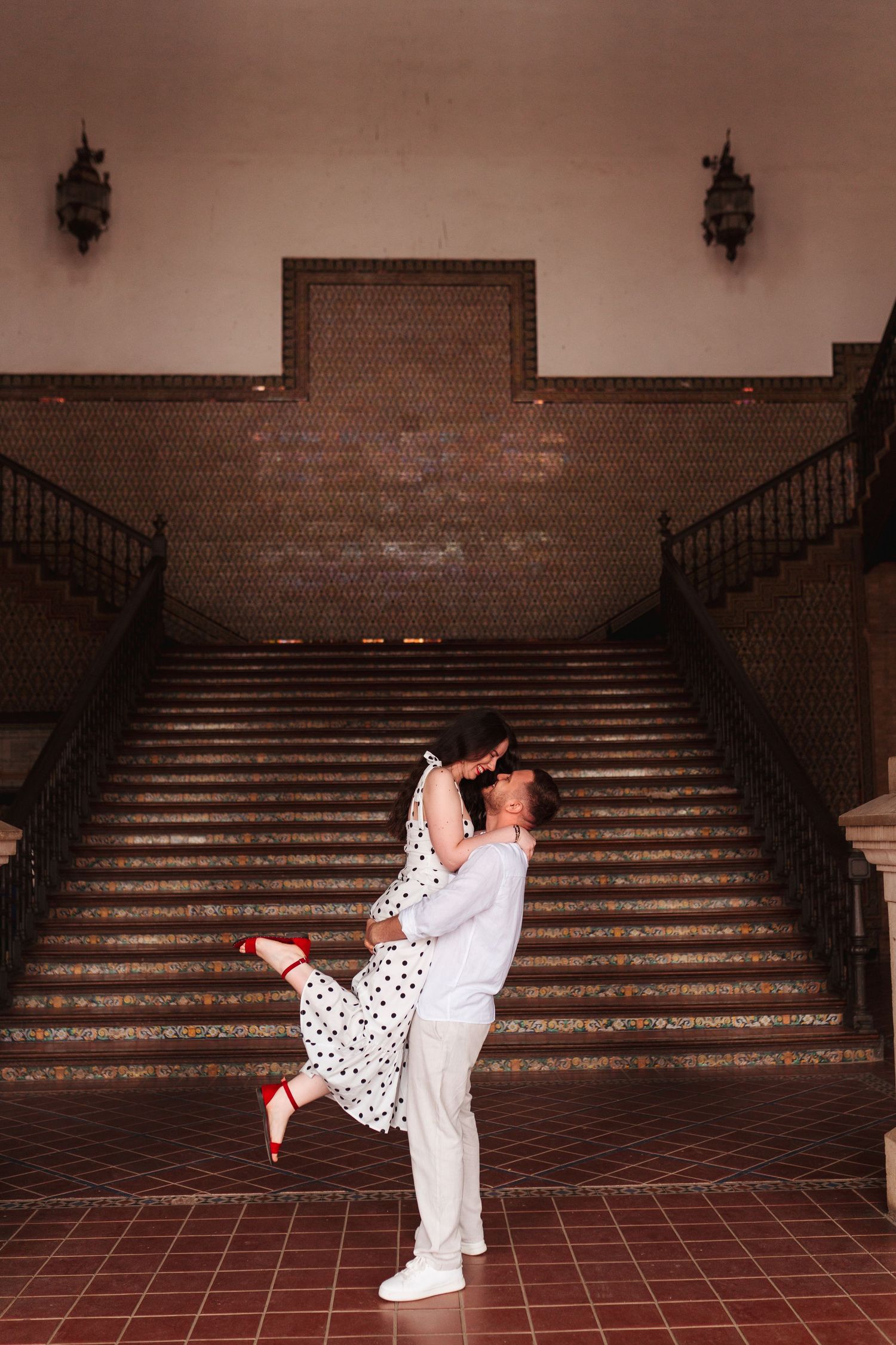 A man lifts a woman in a polka dot dress and red heels on a tiled staircase in a romantic couples photo.