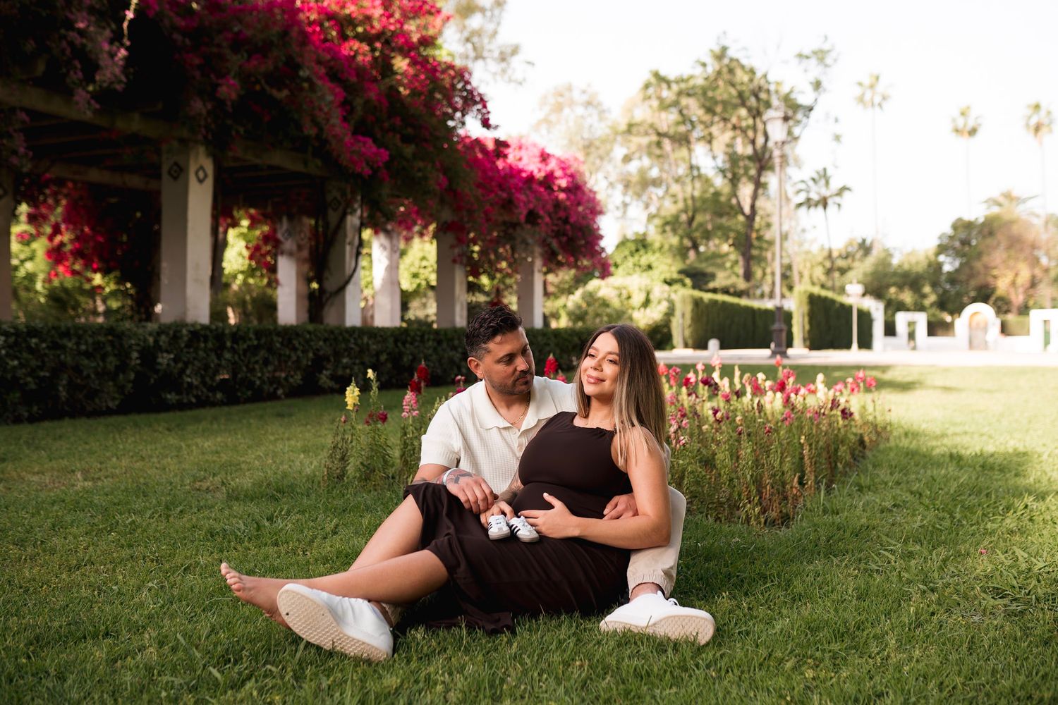 Couple relaxing on grass under pink flowering pergola, woman in black maternity dress holding ultrasound.