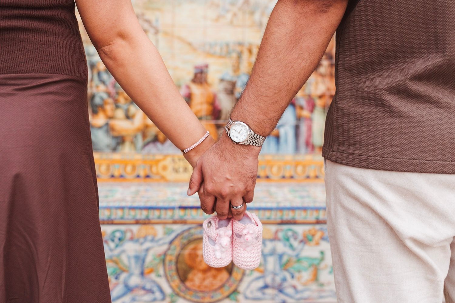 Expecting couple holding hands and tiny pink baby shoes at Plaza de España.