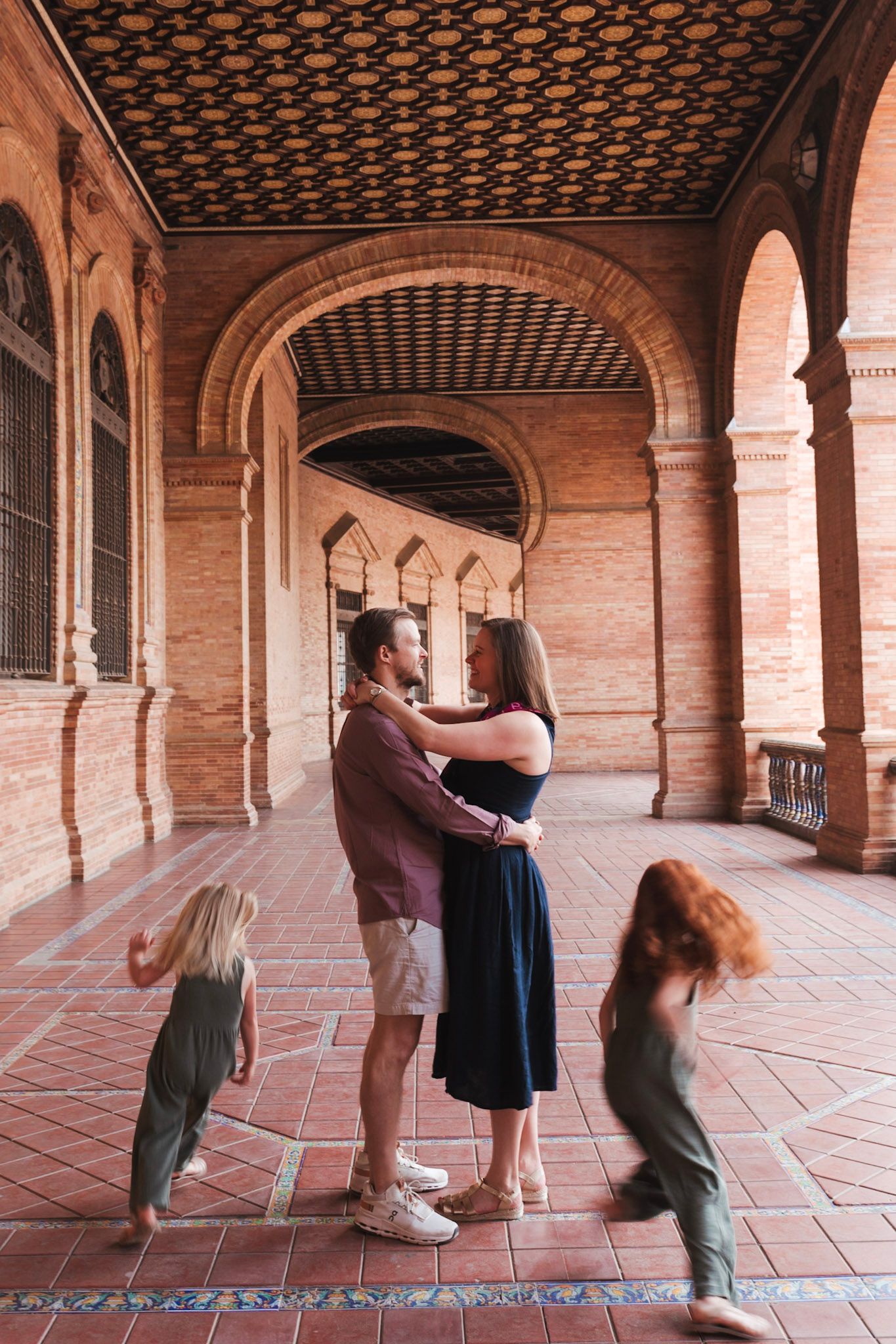 A couple embracing romantically under a brick archway while two children play around them in Seville.