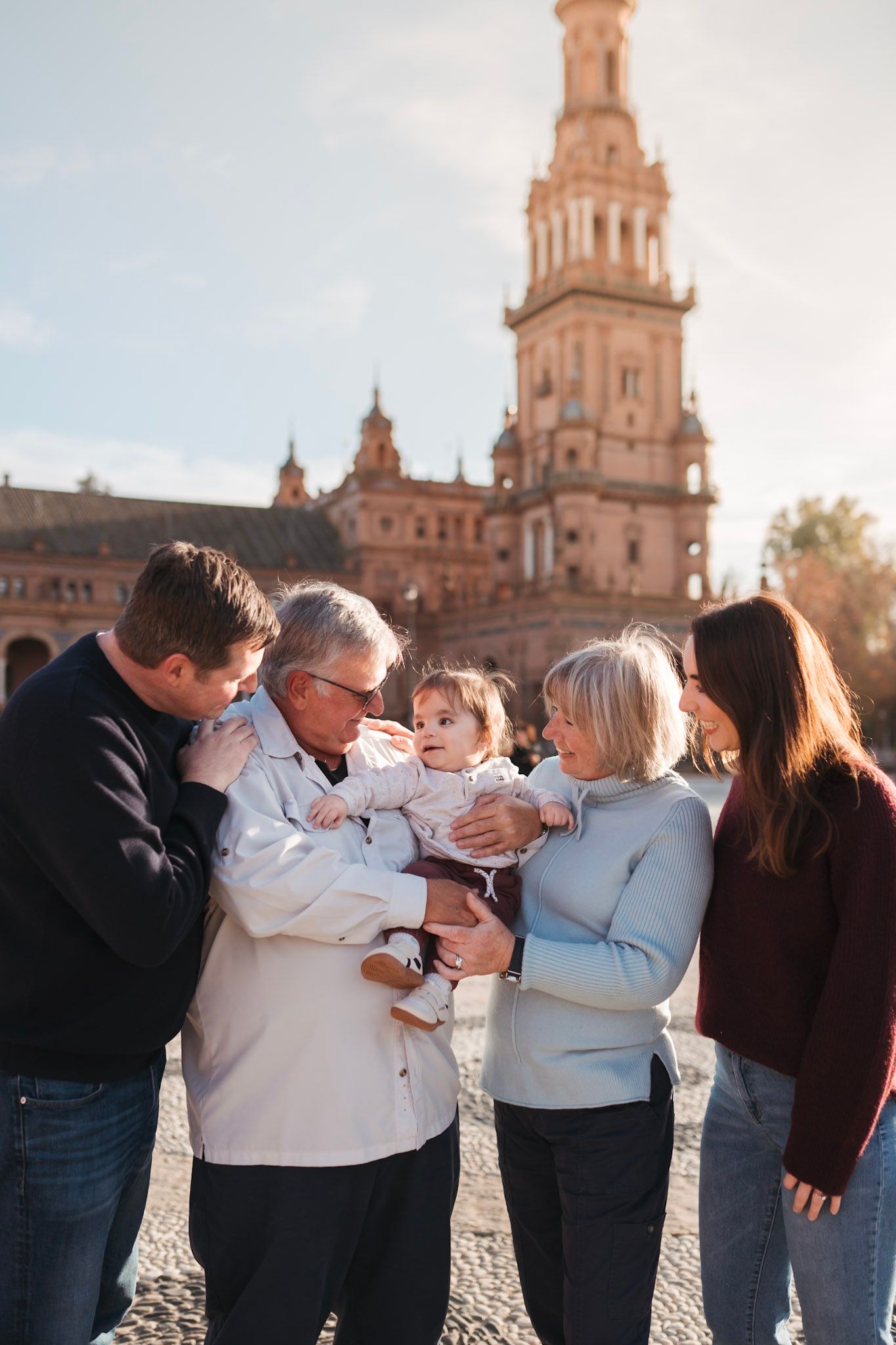 A multigenerational family gathers around a baby at Plaza de España, Seville, on a bright sunny day.