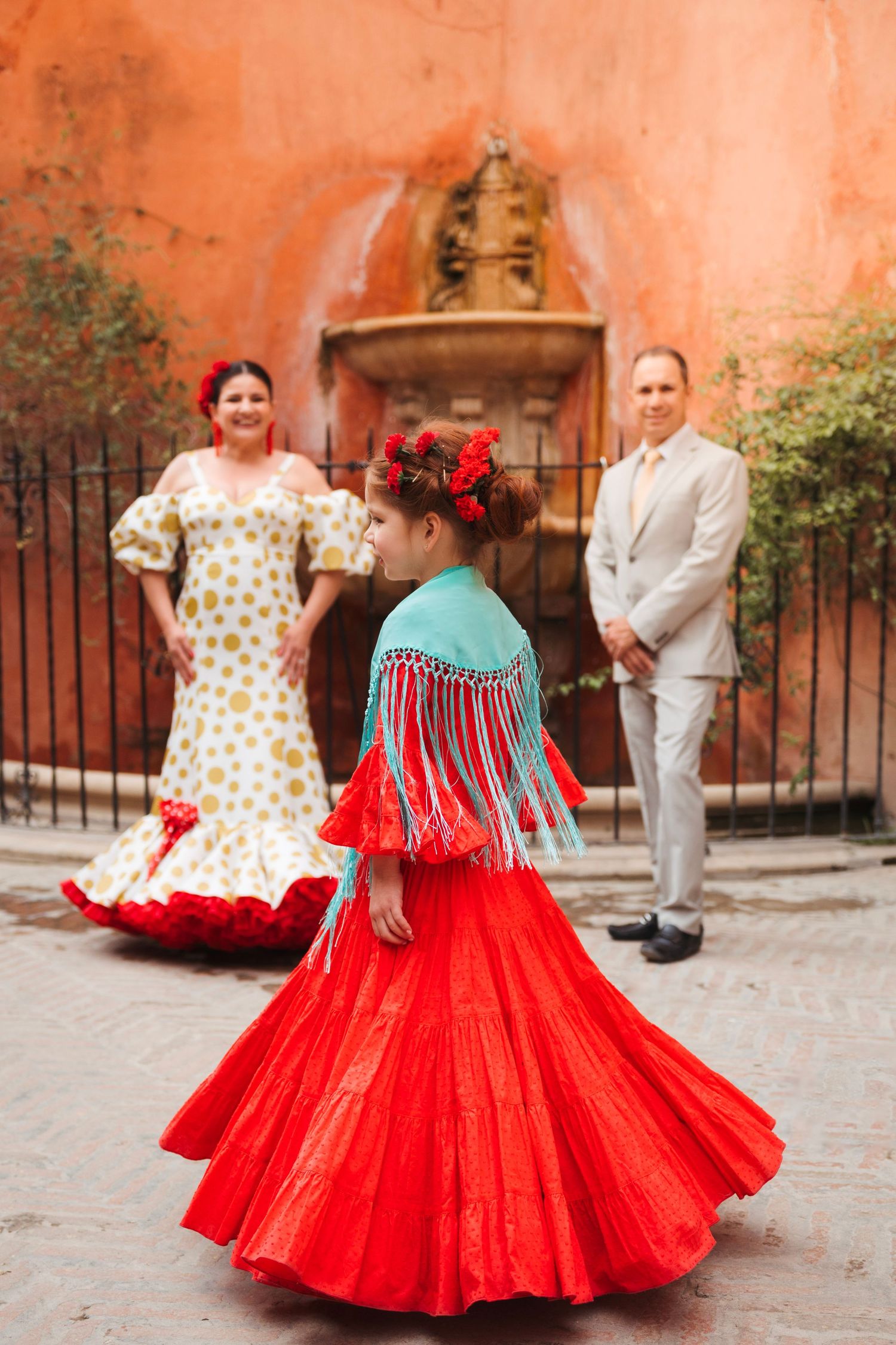 Child in red flamenco dress spins before couple near ornate fountain in Seville.