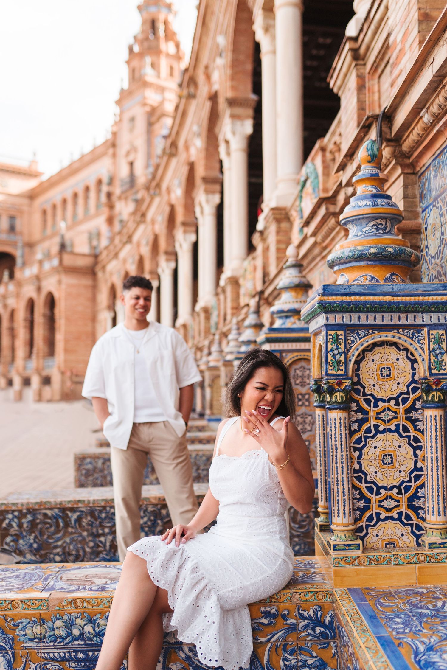 Couple at Plaza de España Seville, woman in white dress sitting on colorful tiled bench, man in white shirt behind her.