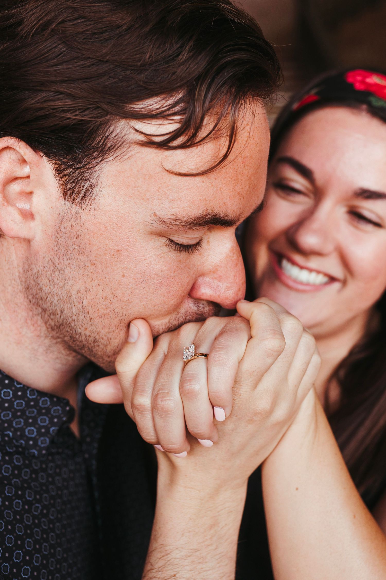 Man kisses woman's hand showing engagement ring as she smiles joyfully in warm indoor setting.