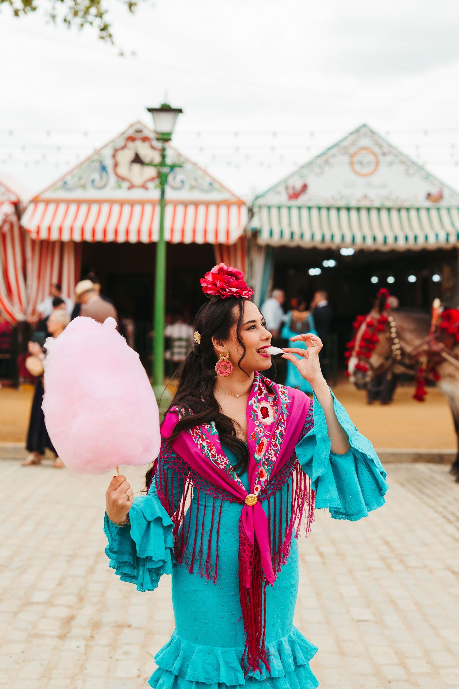 Young woman in turquoise flamenco dress eating pink cotton candy at a Spanish fair.
