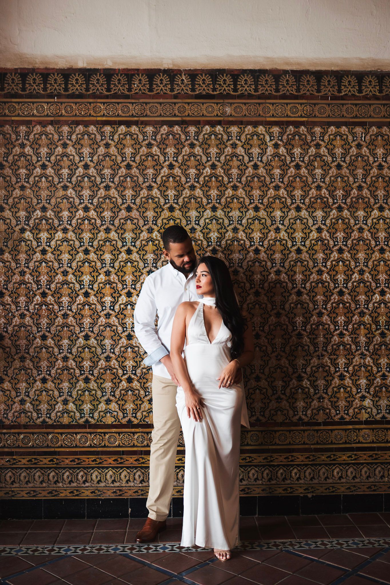 Couple posing against ornate mosaic tile wall, woman in white dress, man in white shirt, elegant portrait.