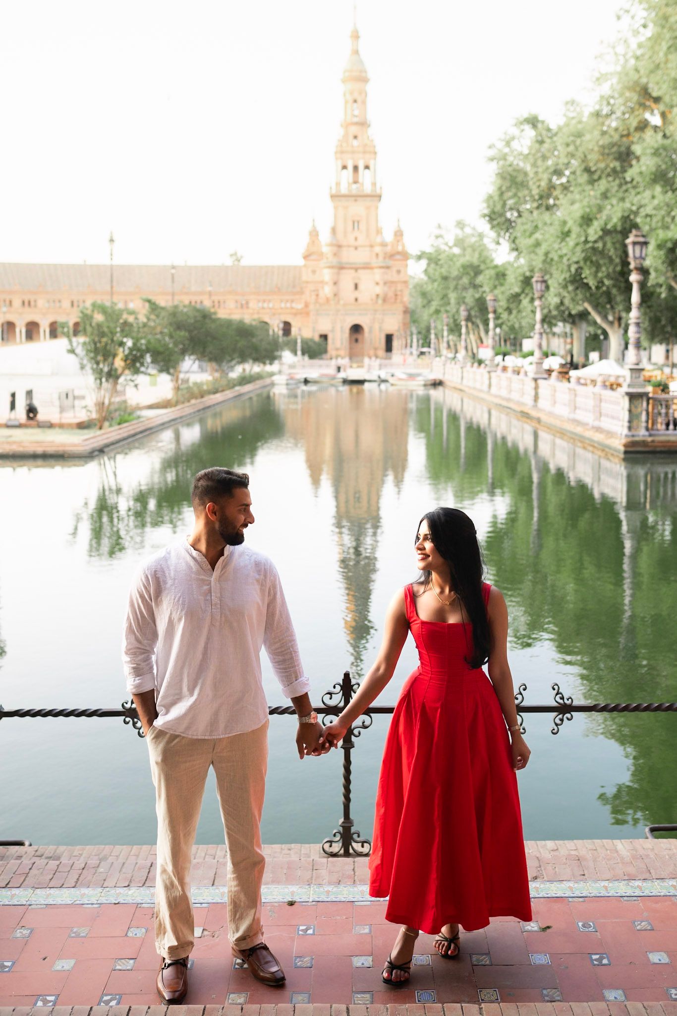 Couple holding hands in a red dress and white shirt by the canal at Plaza de España, Seville, with tower reflection in water.