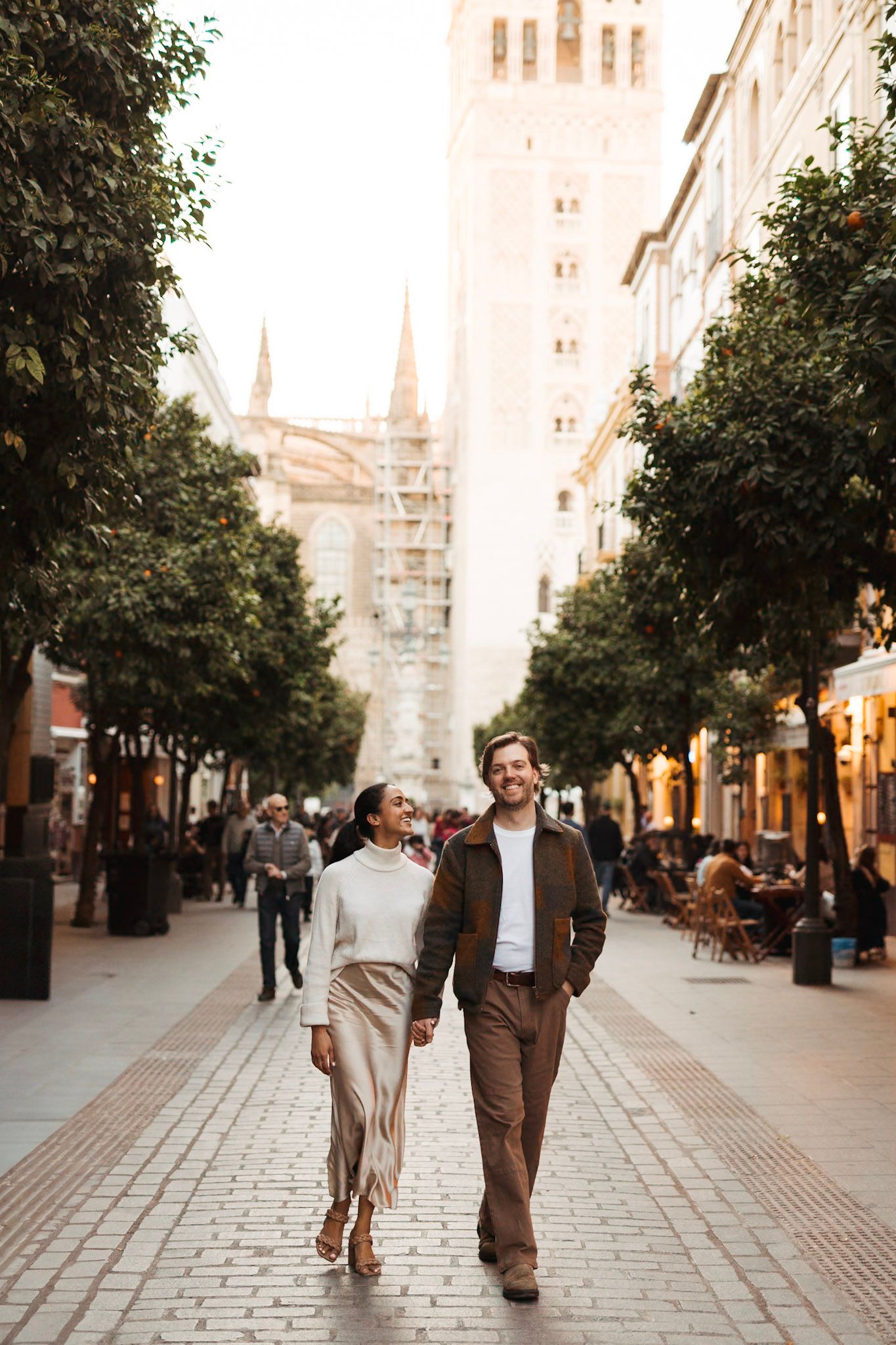 Stylish couple walking hand in hand along a tree-lined street in Seville, Spain.