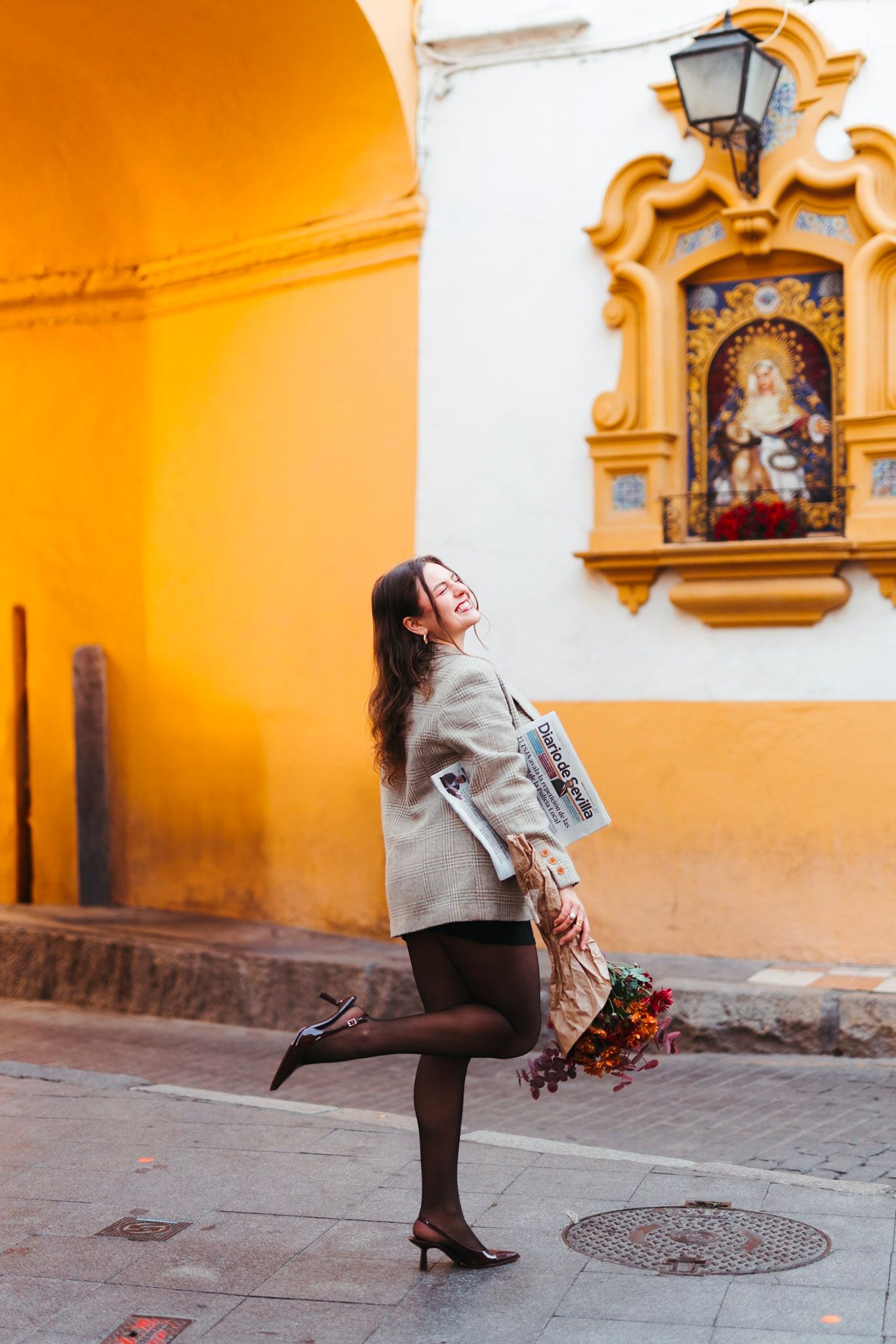 Smiling woman in beige jacket holds flowers while posing on cobblestone street near ornate Seville shrine.